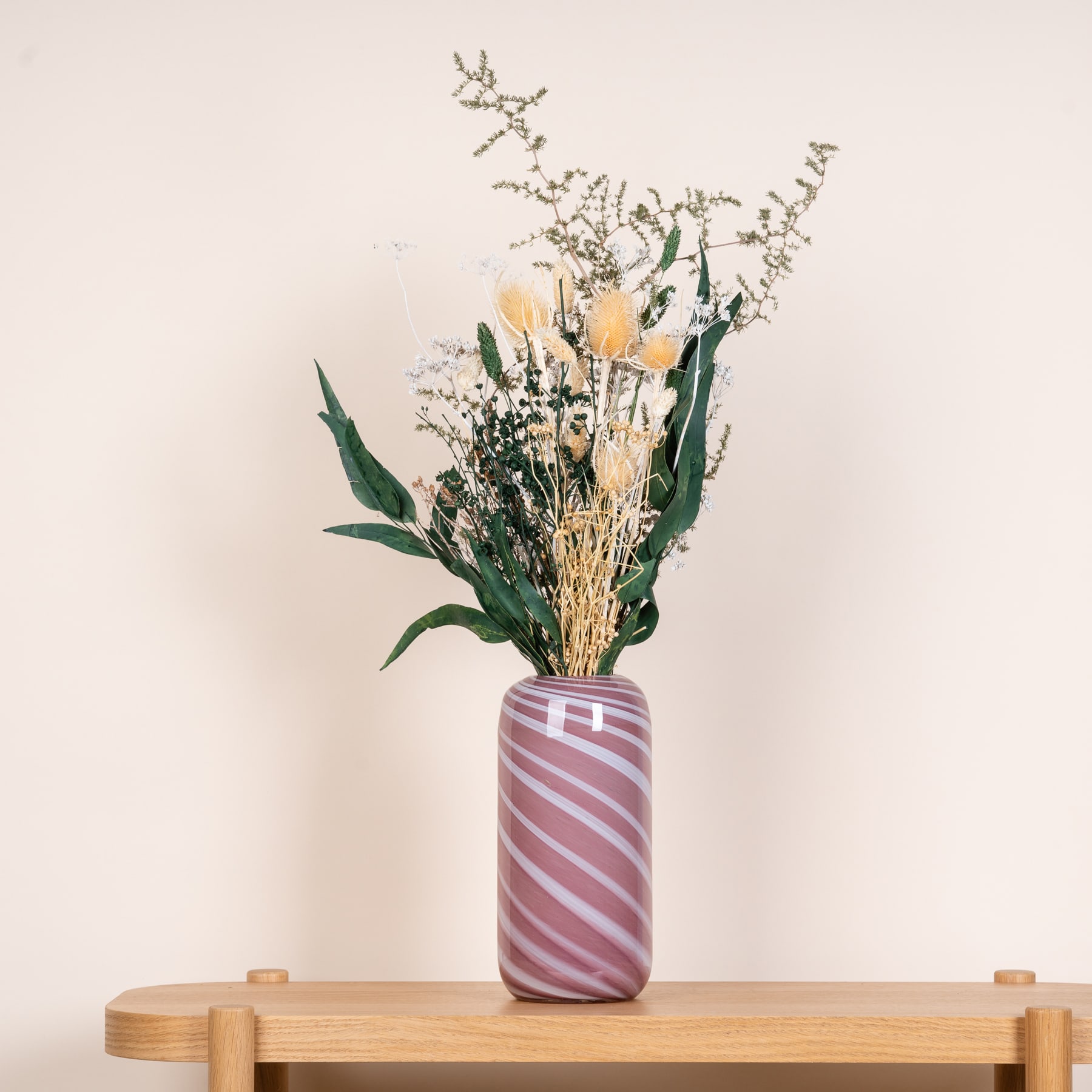 Eine Vase mit einem Strauß getrockneter Blumen und Grünpflanzen steht auf einem Holzregal vor einem schlichten beigen Hintergrund.