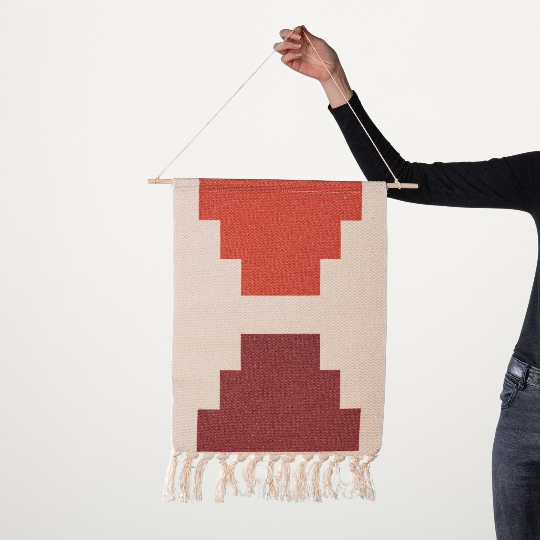 Eine Hand hält ein dekoratives gewebtes Wandbehang mit geometrischen Formen in Rot- und Beigetönen vor einem schlichten Hintergrund.
