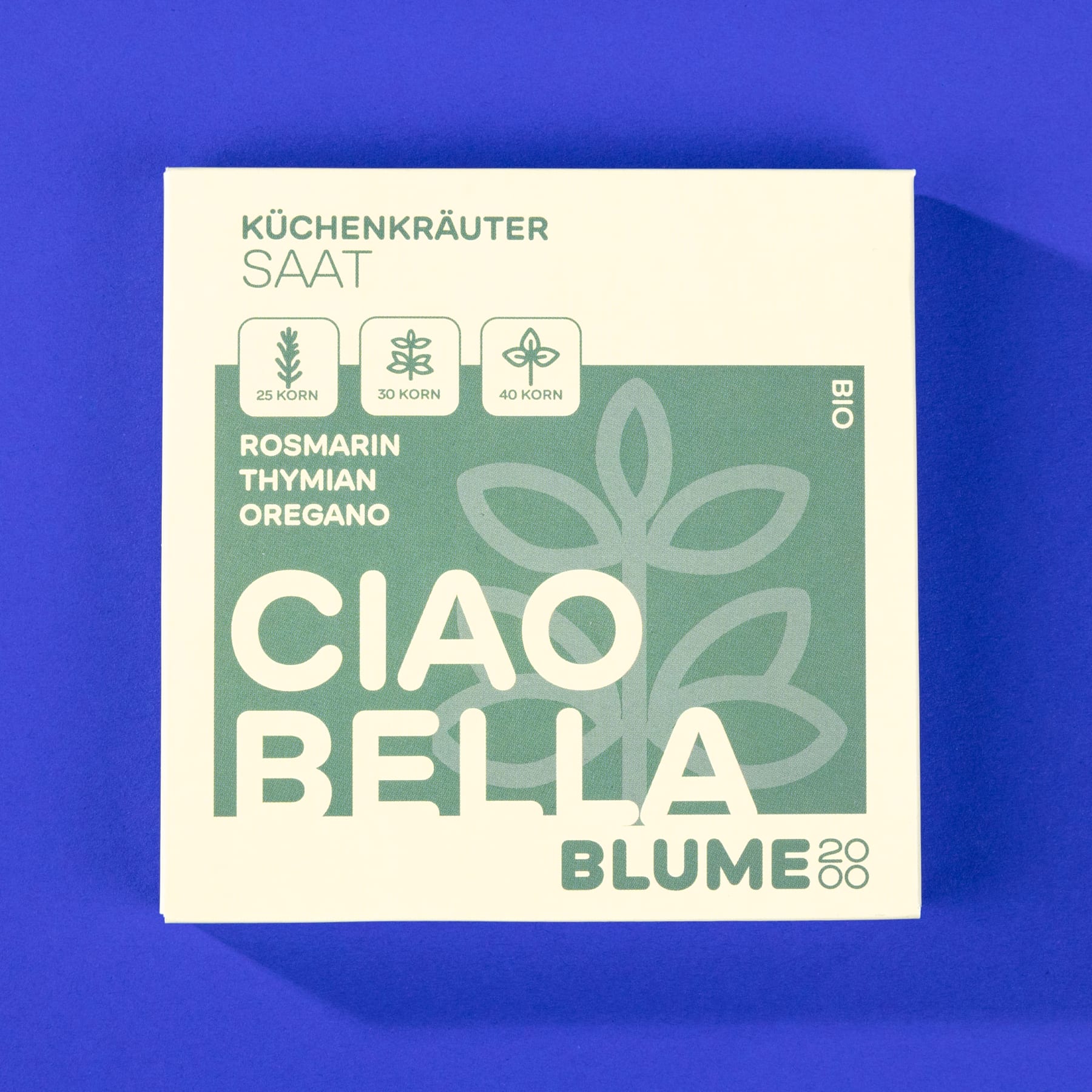 Das Bild zeigt eine grün-weiße quadratische Verpackung mit der Aufschrift „Ciao Bella Blume“ und Symbolen für Rosmarin, Thymian und Oregano vor einem leuchtend blauen Hintergrund.