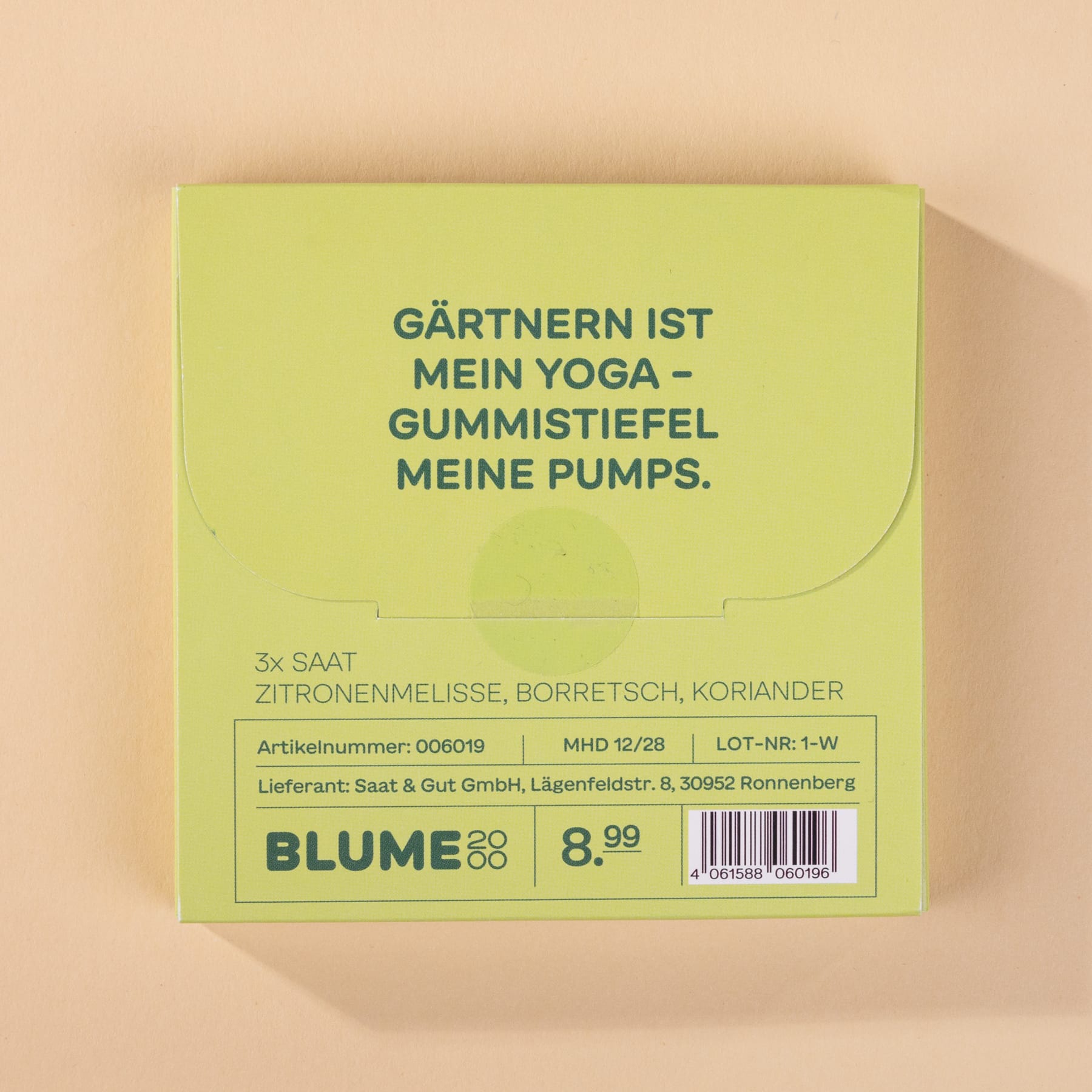 Das Bild zeigt eine grüne Schachtel mit dem deutschen Text „Gärtnern ist mein Yoga – Gummistiefel meine Pumps“. Die Schachtel scheint ein Produkt zu sein, mit einem Strichcode und anderen sichtbaren Produktinformationen.