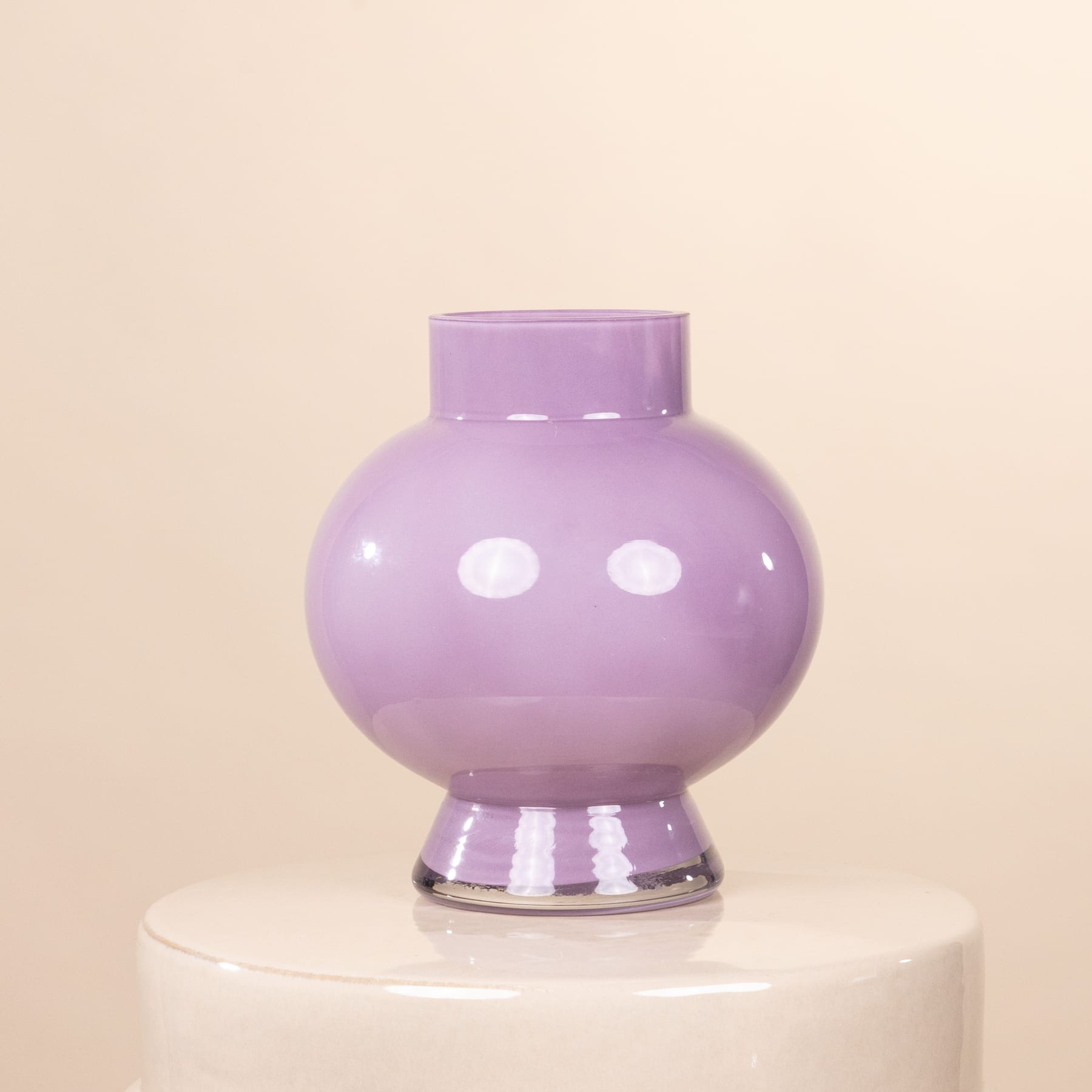 Eine pastellfarbene Vase mit zwei leuchtend rosa Gerbera-Blumen steht auf einem runden Holztisch vor einem schlichten weißen Hintergrund.
