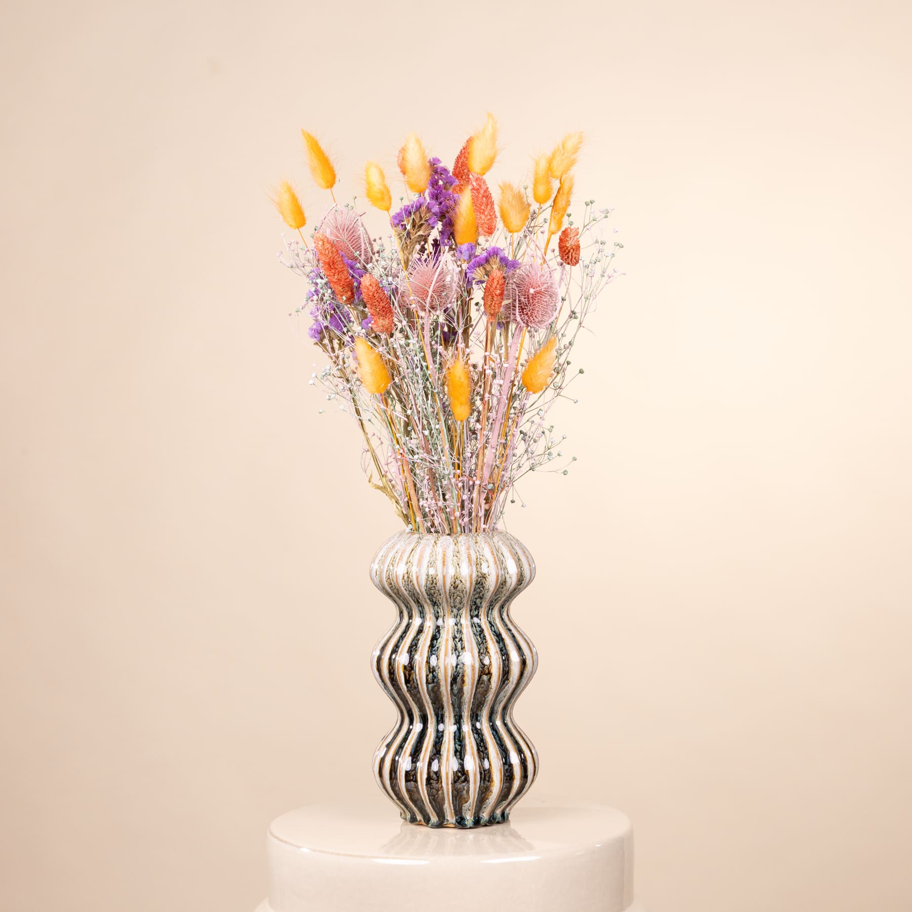 Ein farbenfroher Strauß getrockneter Blumen in einer auffälligen, gewellten Vase steht auf einer schlichten, weißen Oberfläche vor einem weichen, beigen Hintergrund.