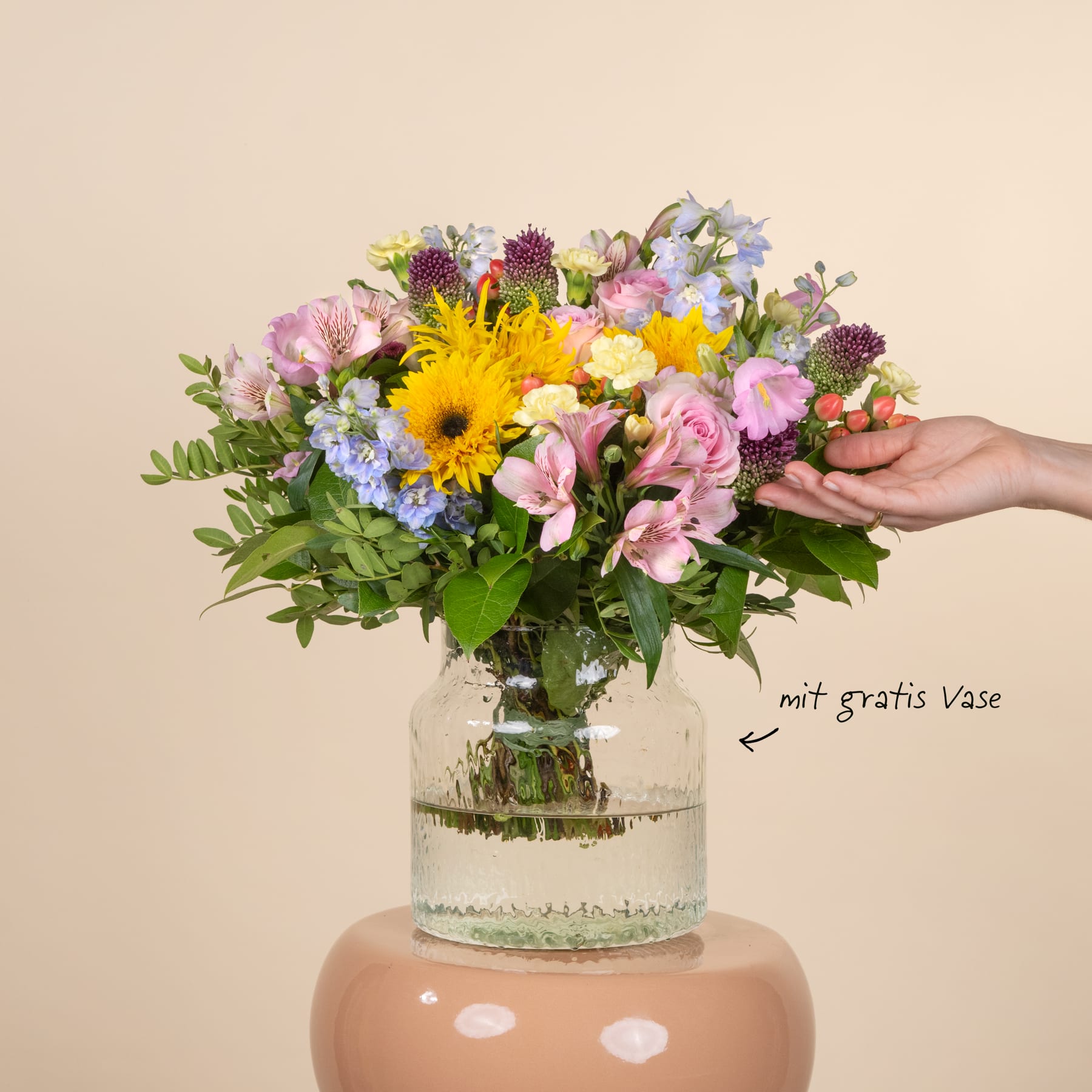 Ein lebendiger Strauß bunter Blumen, darunter Sonnenblumen, Pfingstrosen und andere Blüten, steht in einer Glasvase vor einem schlichten beigen Hintergrund.