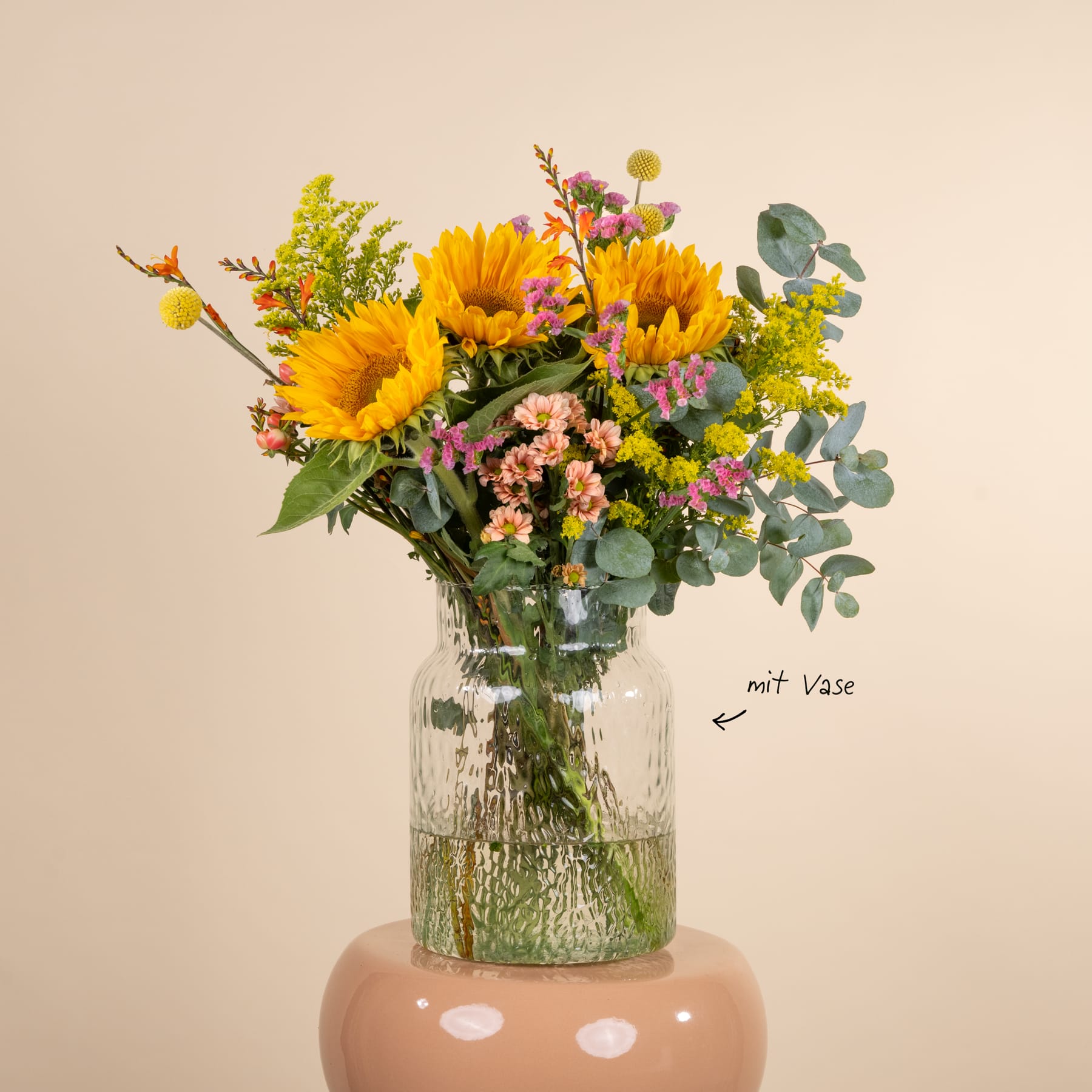 Ein farbenfroher Strauß aus Sonnenblumen, Wildblumen und Grünpflanzen in einer Glasvase steht auf einem schlichten beigen Hintergrund.