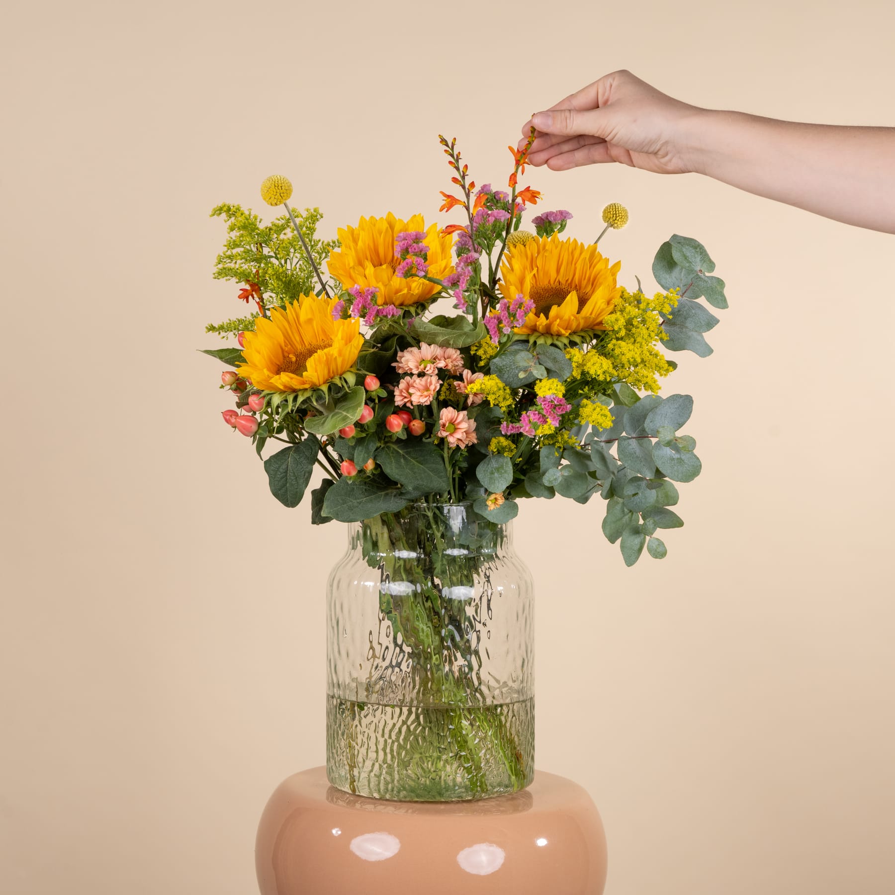 Ein lebendiger Strauß aus Sonnenblumen, Gänseblümchen und anderen bunten Blumen, arrangiert in einer Glasvase vor einem neutralen beigen Hintergrund.