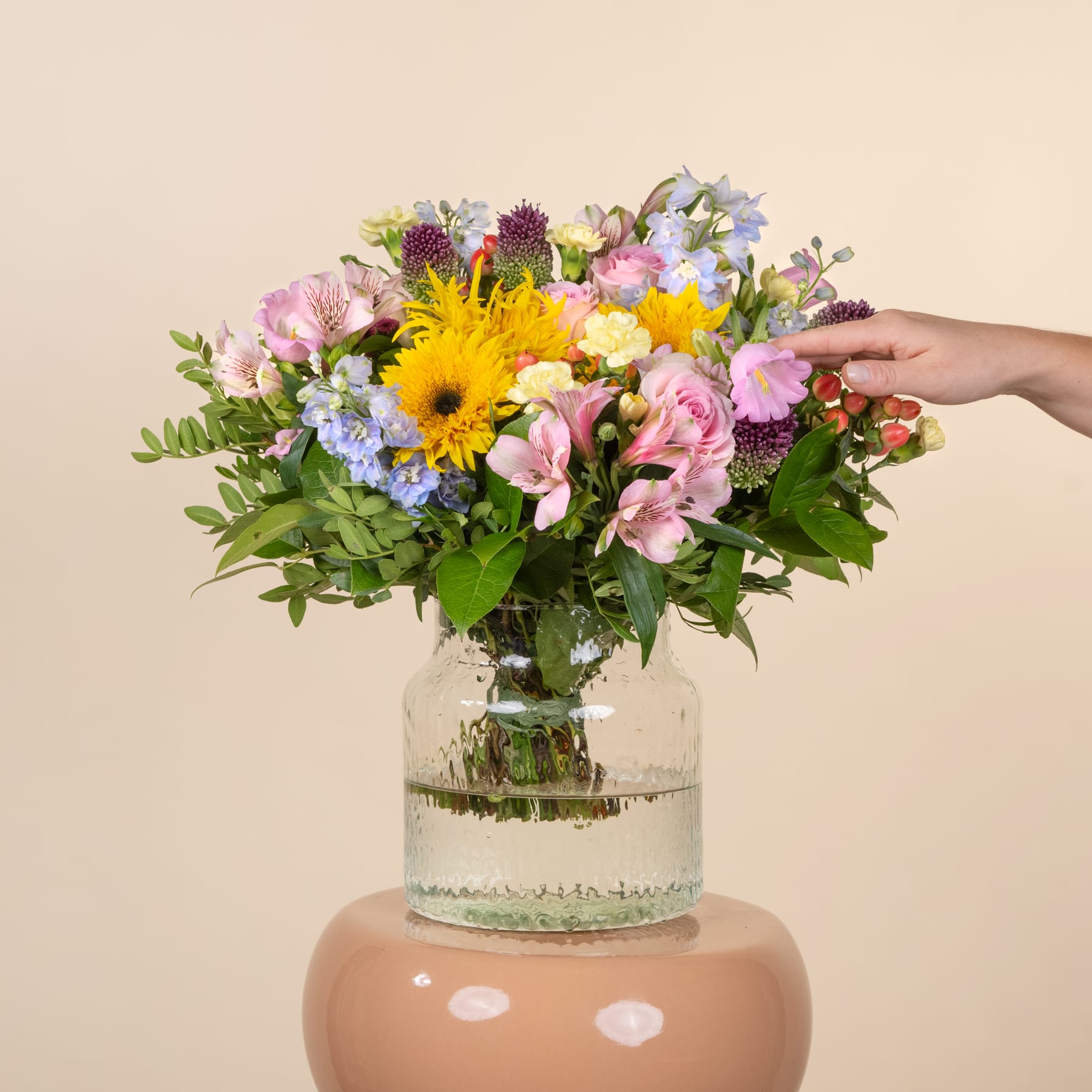 Ein lebendiger Strauß bunter Blumen, darunter Sonnenblumen, Pfingstrosen und andere Blüten, wird in einer Glasvase vor einem schlichten, beigen Hintergrund präsentiert.