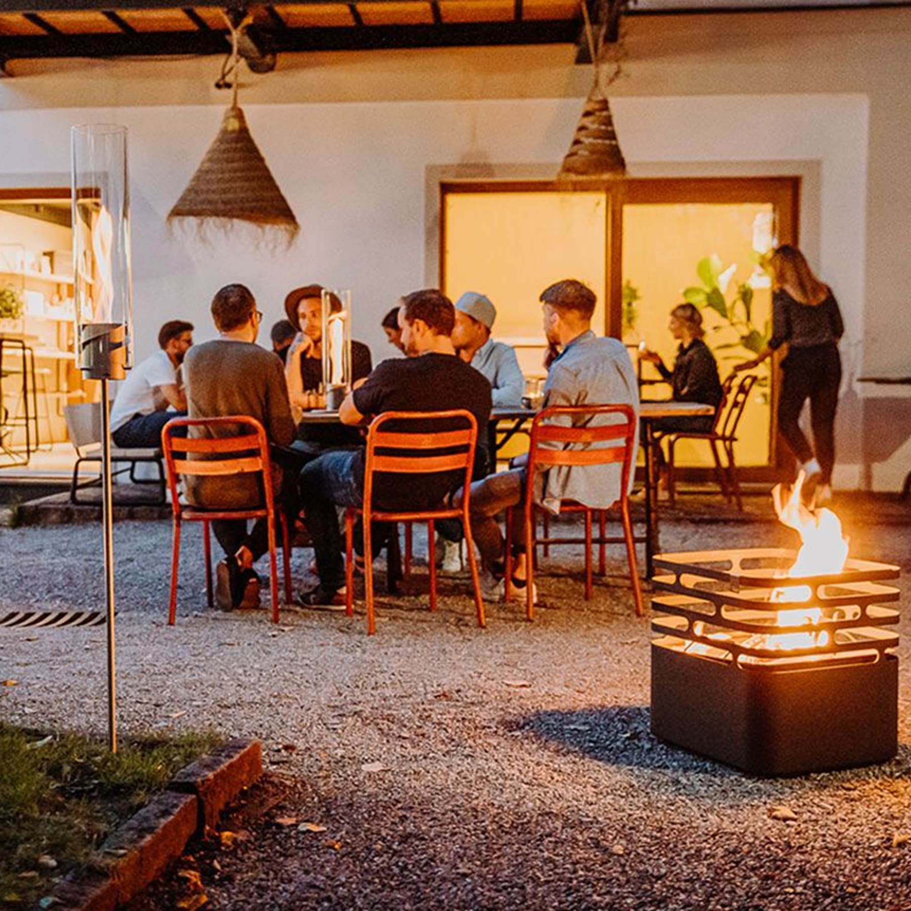 Eine Gruppe von Menschen versammelt sich um einen Tisch im Freien, mit einer Feuerstelle im Vordergrund und einer rustikalen, gemütlichen Atmosphäre im Hintergrund.