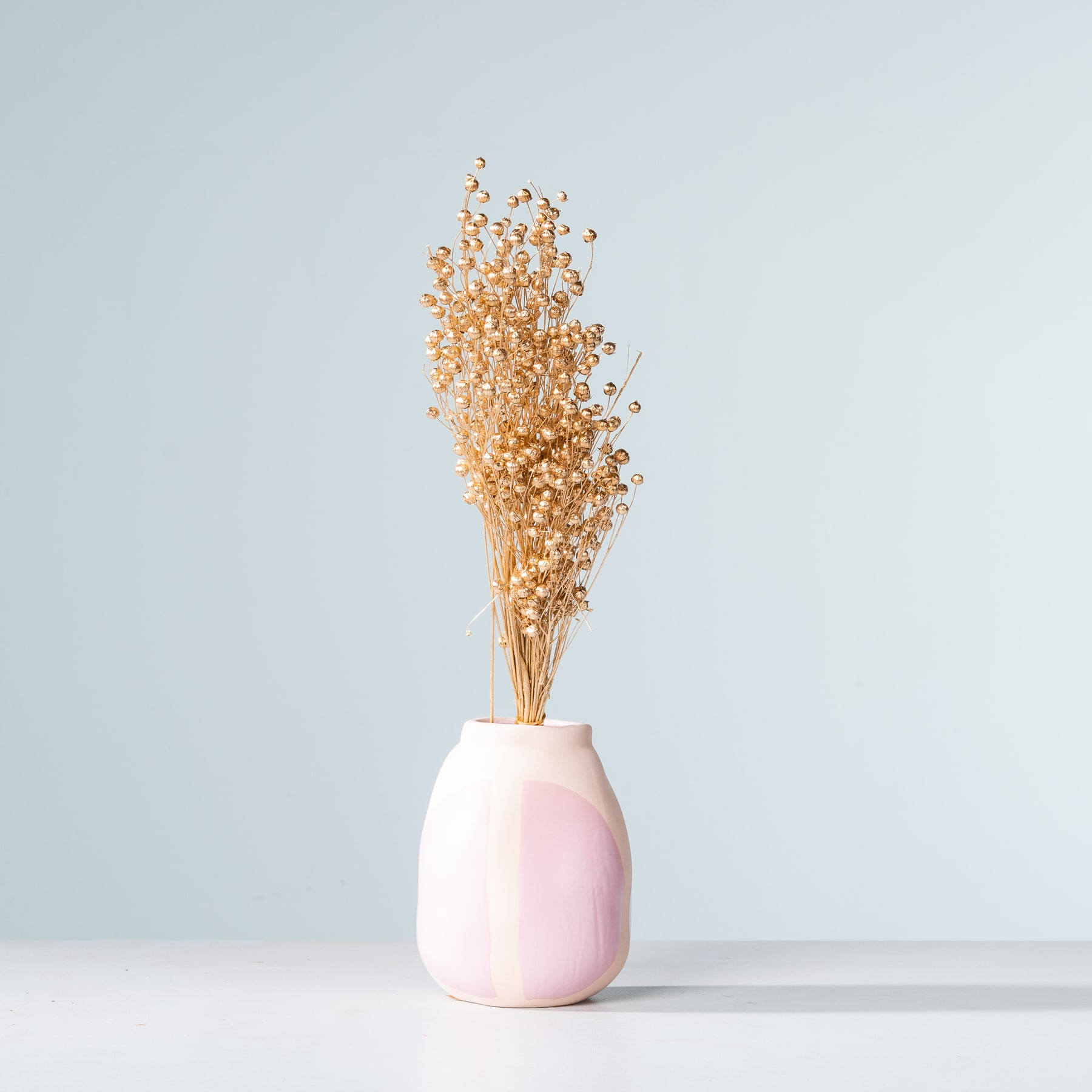 Ein zartes Arrangement aus getrockneten Blumen in einer zartrosa Vase vor einem hellblauen Hintergrund.