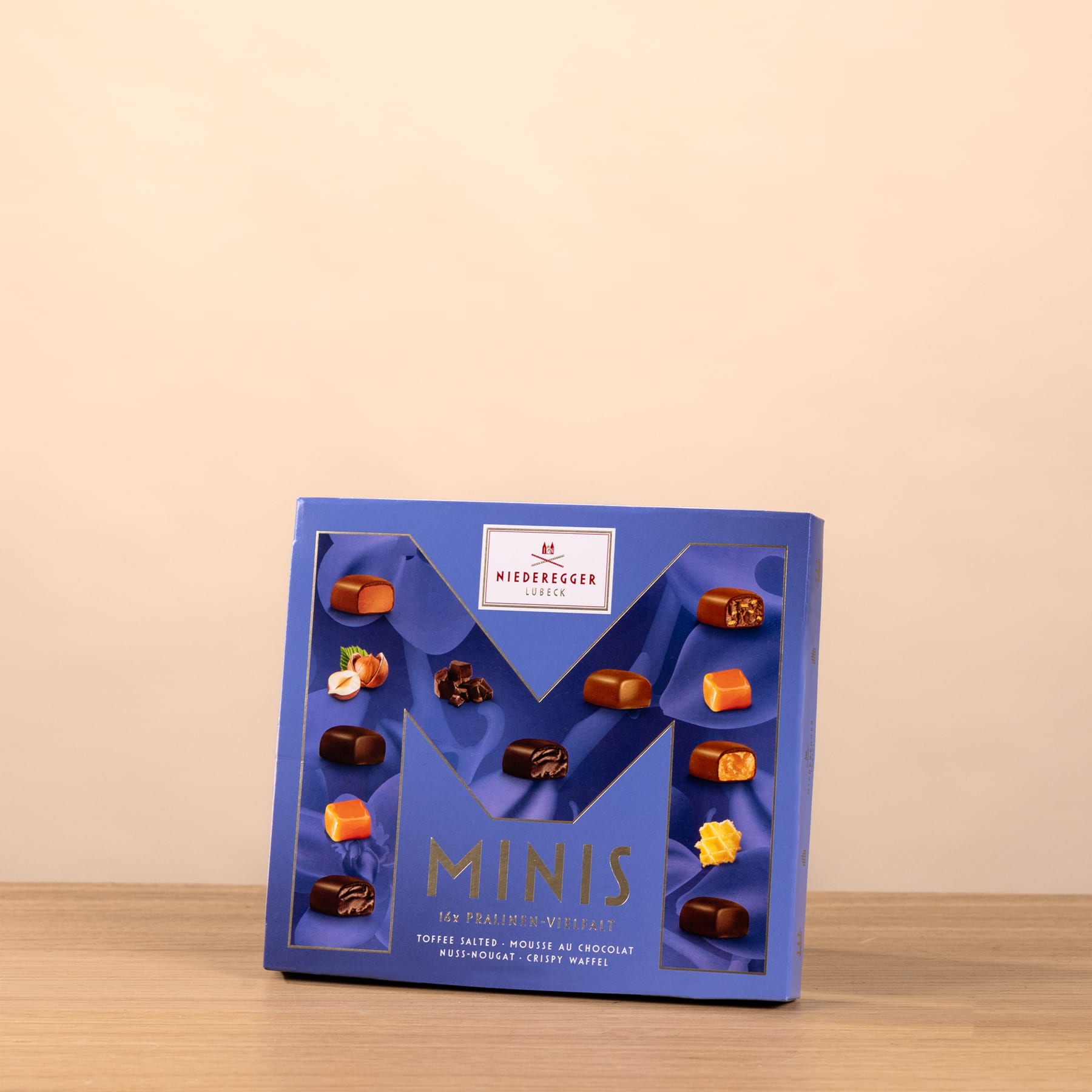 Eine blaue Schachtel mit verschiedenen Schokoladenbonbons steht auf einer Holzoberfläche vor einem hellpfirsichfarbenen Hintergrund.