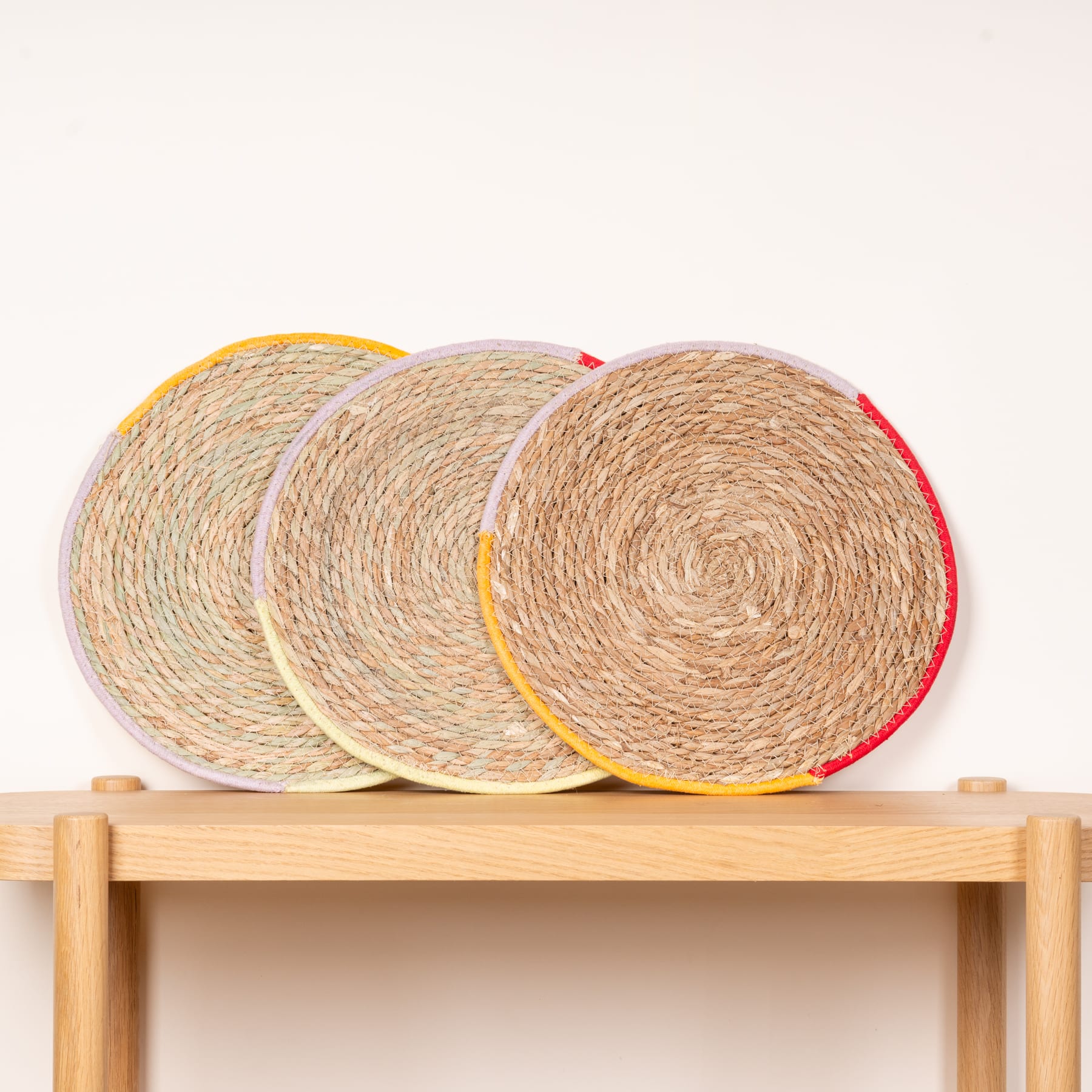 Drei runde gewebte Tischsets oder Untersetzer aus Naturfasern werden auf einem Holzregal vor einem schlichten weißen Hintergrund präsentiert.