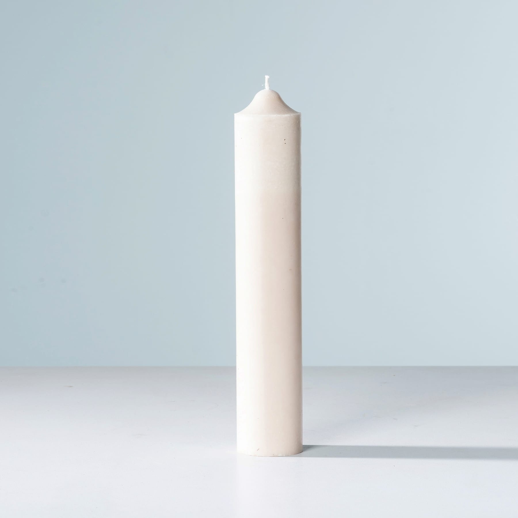 Eine große, weiße Kerze steht allein auf einer schlichten, weißen Oberfläche vor einem hellblauen Hintergrund.