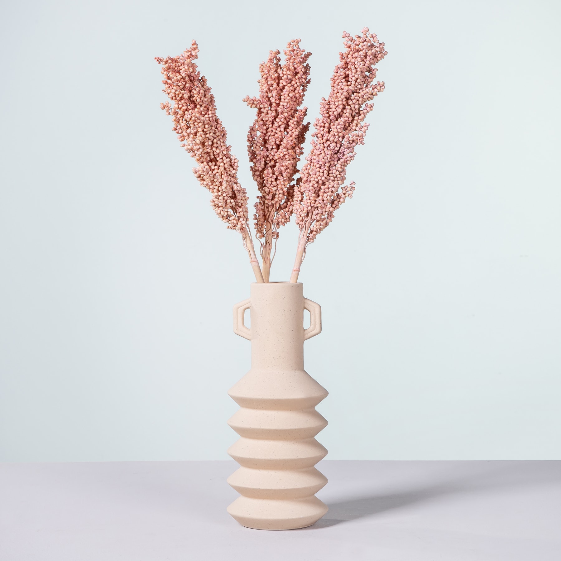 Eine blassrosa Keramikvase mit Spiralmuster hält mehrere zarte, rosafarbene Blumenstiele vor einem schlichten, hellen Hintergrund.