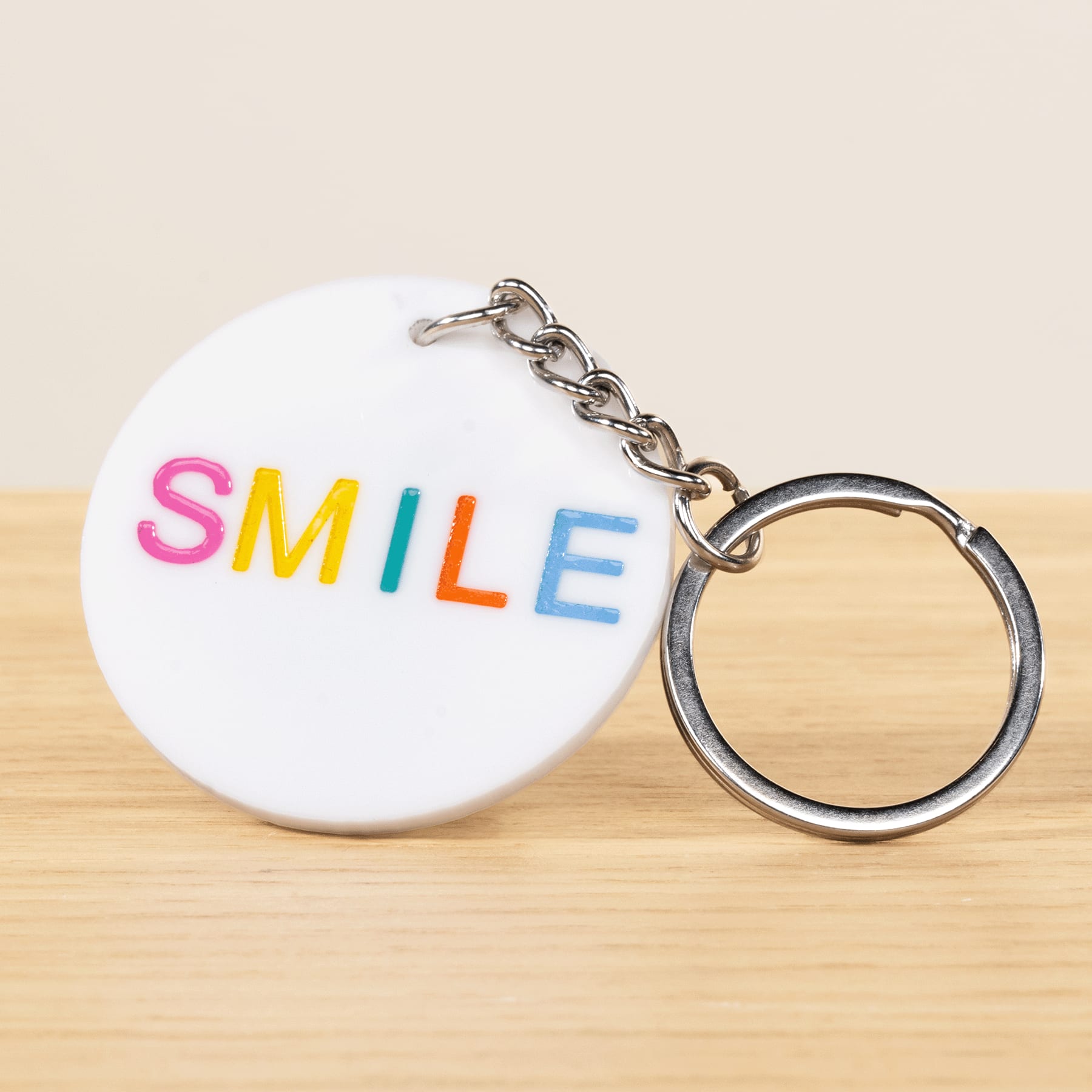 Auf einer Holzoberfläche liegt ein weißer, runder Schlüsselanhänger mit dem Wort „SMILE“ in bunten Buchstaben.