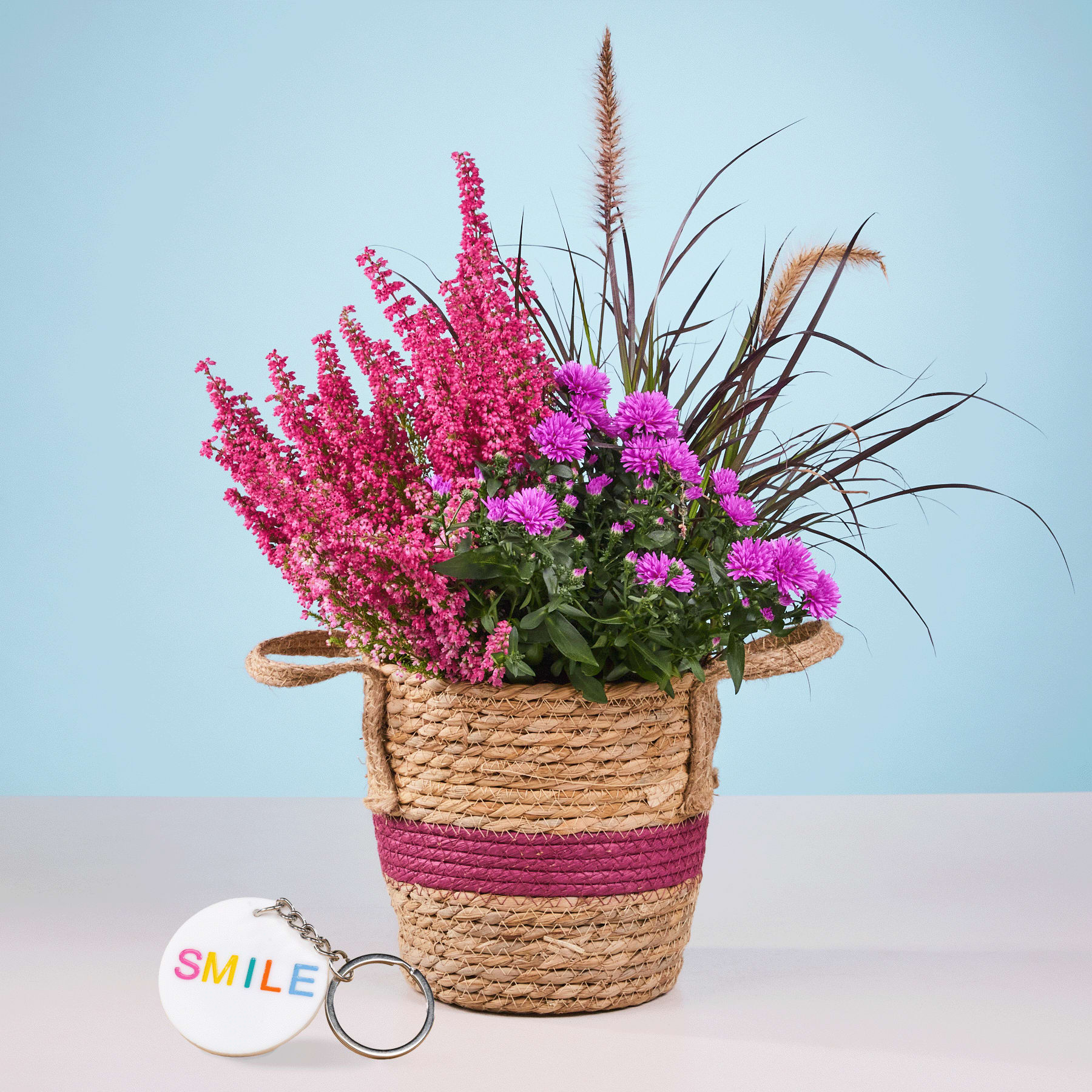 Auf einem hellblauen Hintergrund steht ein geflochtener Korb voller leuchtend rosa und violetter Blumen, darunter hohe Blütenähren und kleinere.