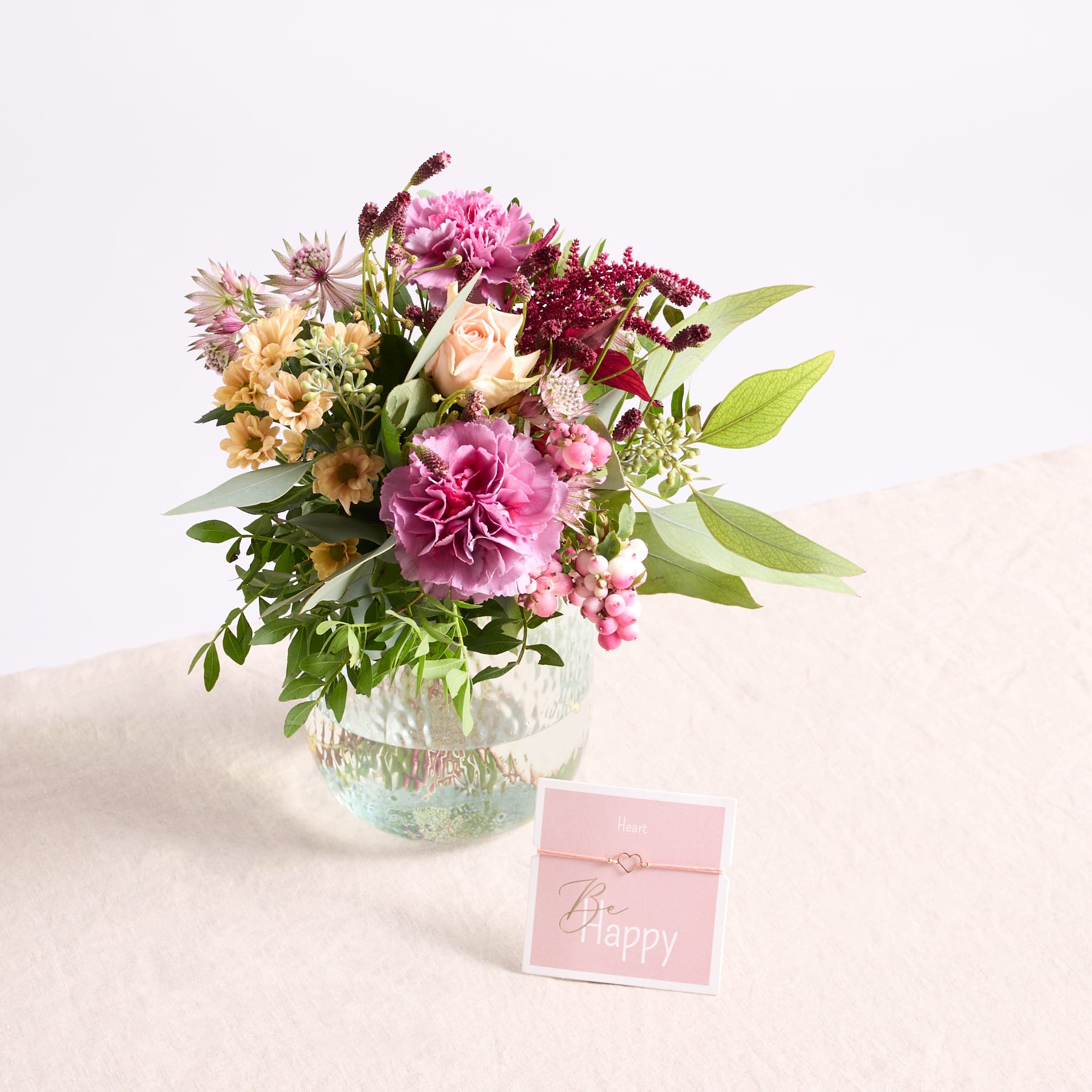 Ein leuchtender Strauß rosa Pfingstrosen, gelben Sonnenblumen und anderen bunten Blumen in einer Glasvase steht auf einer hellen Oberfläche, begleitet von einer kleinen rosa Geschenkbox.