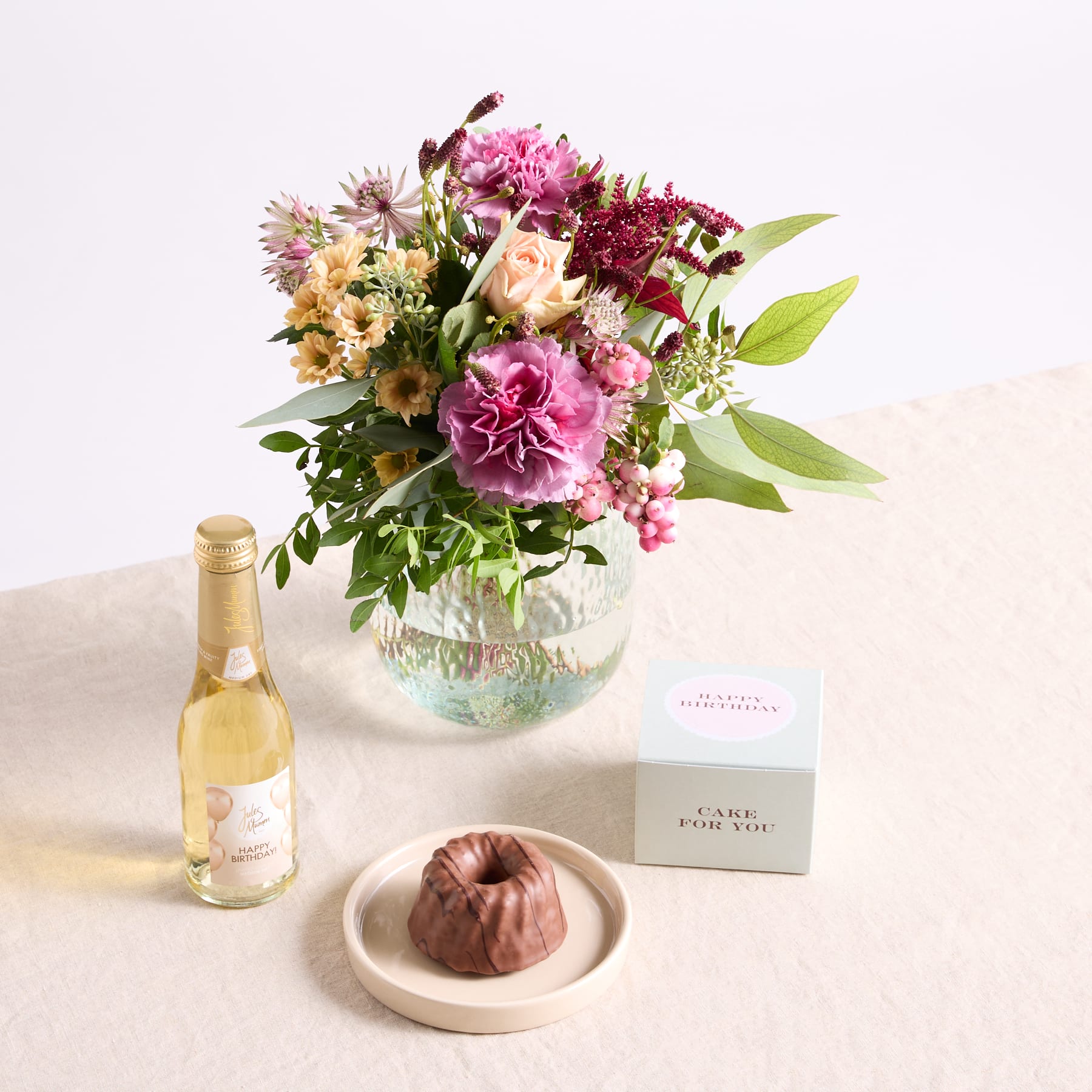 Auf einem Tisch steht ein farbenfroher Strauß rosa und violetter Blumen in einer Glasvase, dazu eine Flasche Wein und ein kleines Dessert auf einem Teller.