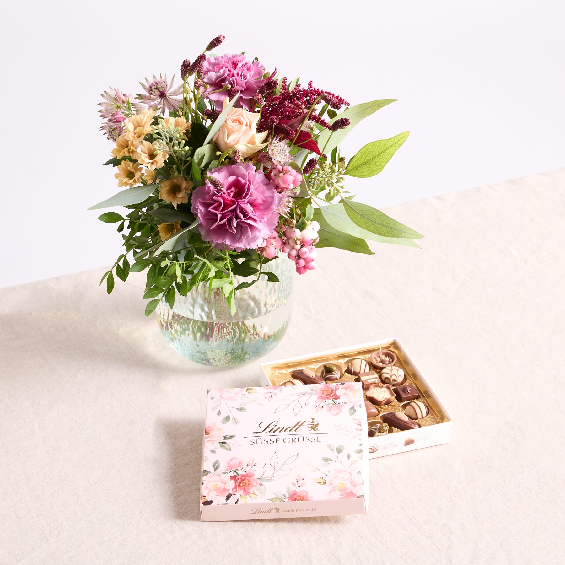 Auf einem Tisch steht ein farbenfroher Strauß rosa und gelber Blumen in einer Glasvase, begleitet von einer Schachtel Lindt-Pralinen.