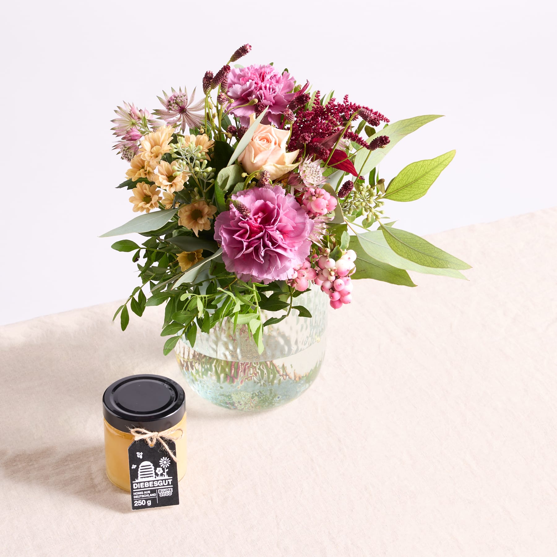 Ein leuchtender Strauß bunter Blumen, darunter rosa Pfingstrosen, gelbe Gänseblümchen und violette Chrysanthemen, wird in einer Glasvase auf einer hellen Oberfläche präsentiert, im Vordergrund steht ein kleines schwarzes Gefäß.