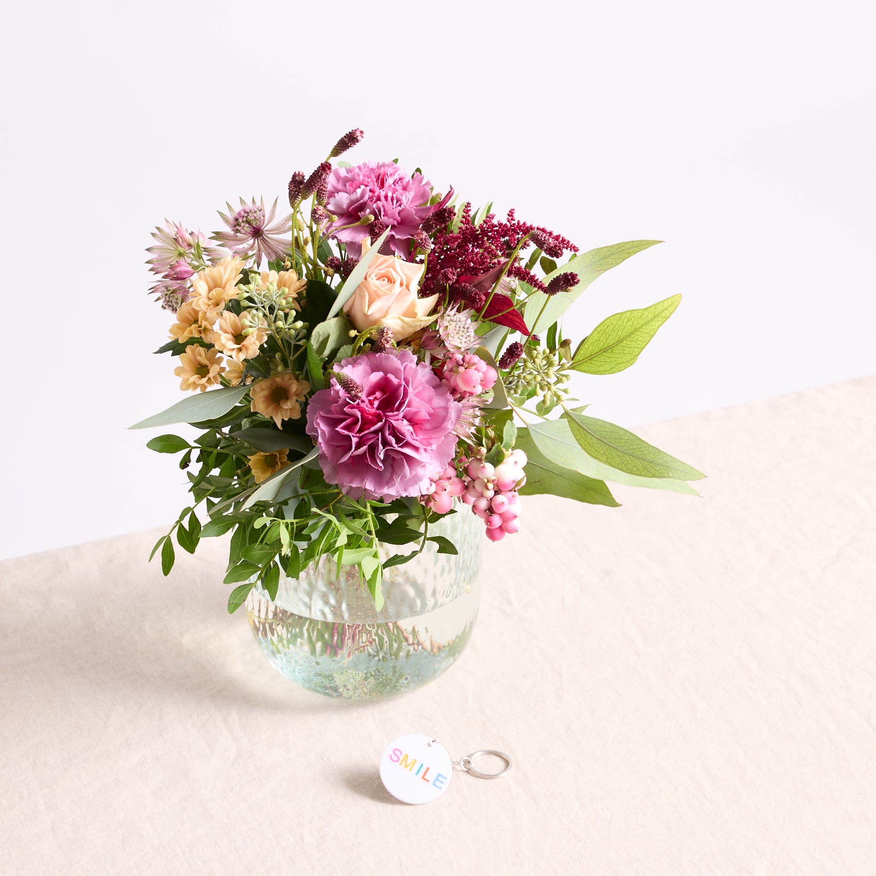 Ein lebendiger Strauß bunter Blumen, darunter rosa Nelken, gelbe Sonnenblumen und grünes Laub, wird in einer Glasvase auf einer hellen Oberfläche präsentiert.