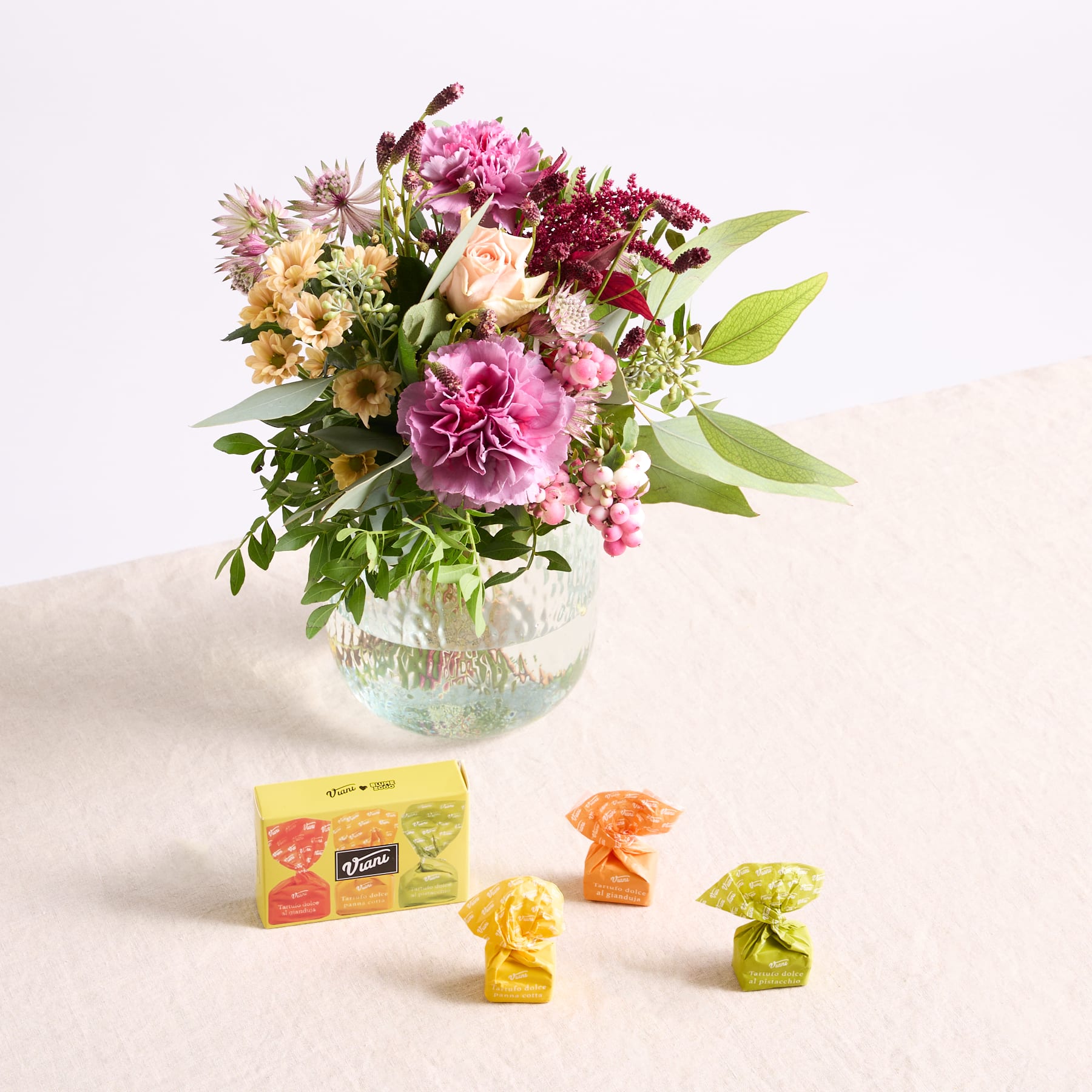 Ein leuchtender Strauß bunter Blumen, darunter Pfingstrosen und andere Blüten, steht in einer Glasvase auf einem Tisch, im Vordergrund stehen eine Schachtel Pralinen und einzeln verpackte Pralinen.