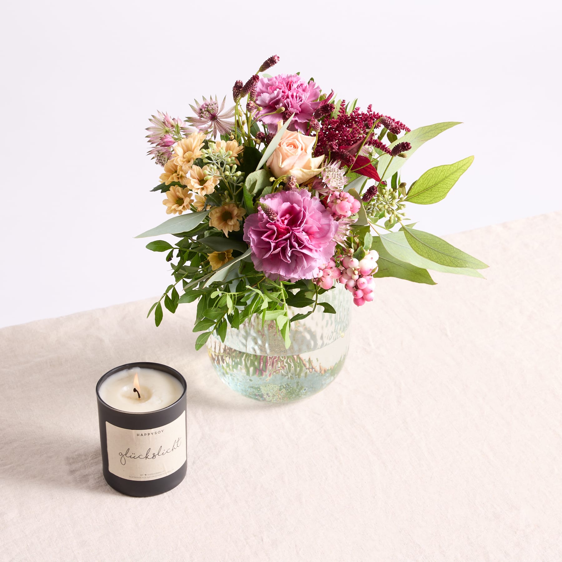Ein leuchtender Strauß bunter Blumen, darunter rosa Pfingstrosen, gelbe Sonnenblumen und violette Chrysanthemen, steht in einer Glasvase auf einer hellen Oberfläche, begleitet von einer schwarzen Kerze.