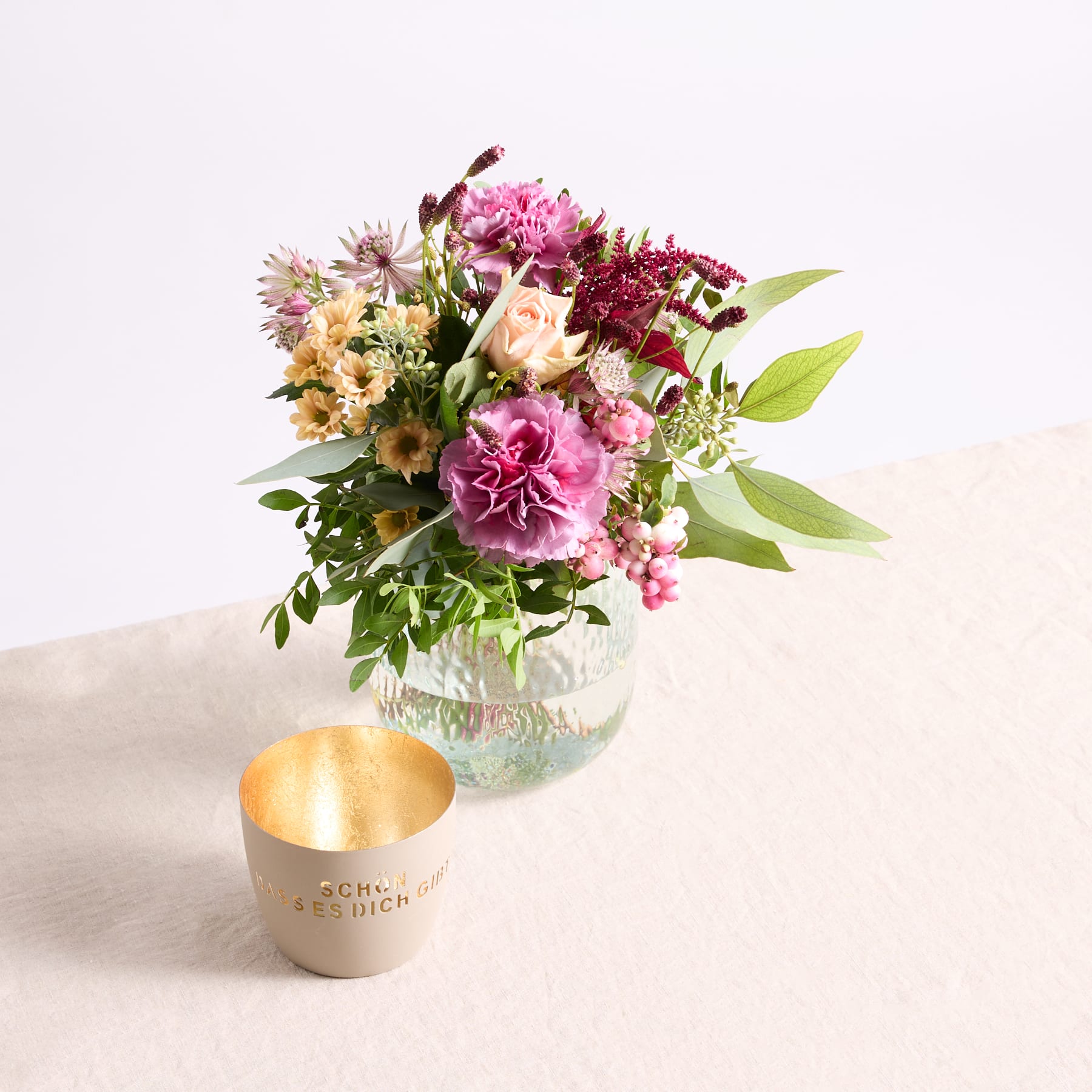 Ein leuchtender Strauß rosa und gelber Blumen in einer Glasvase steht auf einer hellen Oberfläche, begleitet von einem goldfarbenen Kerzenhalter.