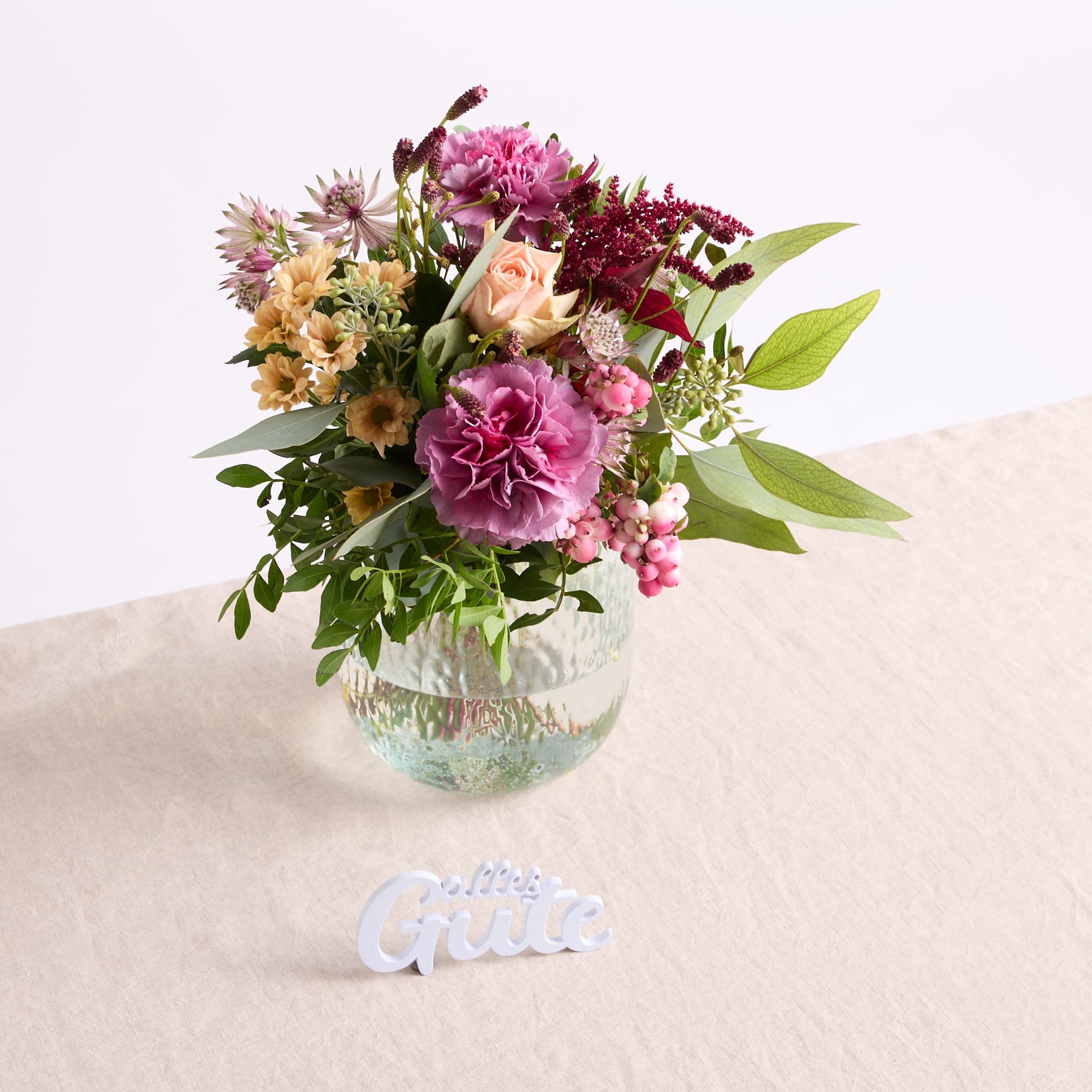 Ein farbenfroher Strauß rosa, gelber und weißer Blumen steht in einer Glasvase auf einer beigen Oberfläche, im Vordergrund ist das Wort „Giver“ zu sehen.