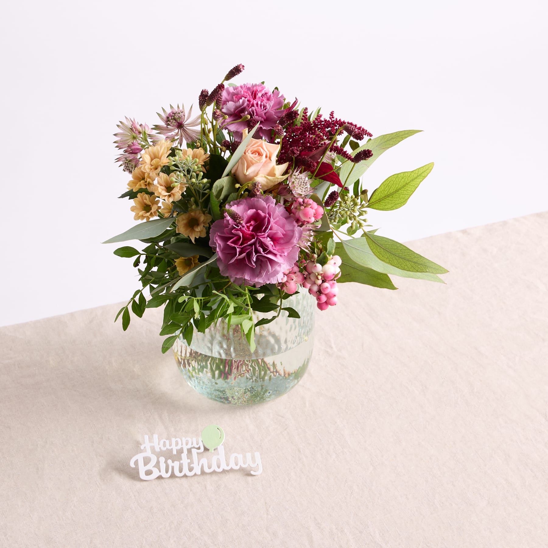 Ein leuchtender Strauß bunter Blumen, darunter rosa Pfingstrosen, Gänseblümchen und andere Blüten, wird in einer Glasvase auf einer hellen Oberfläche präsentiert, im Vordergrund ist die Nachricht „Alles Gute zum Geburtstag“ zu sehen.