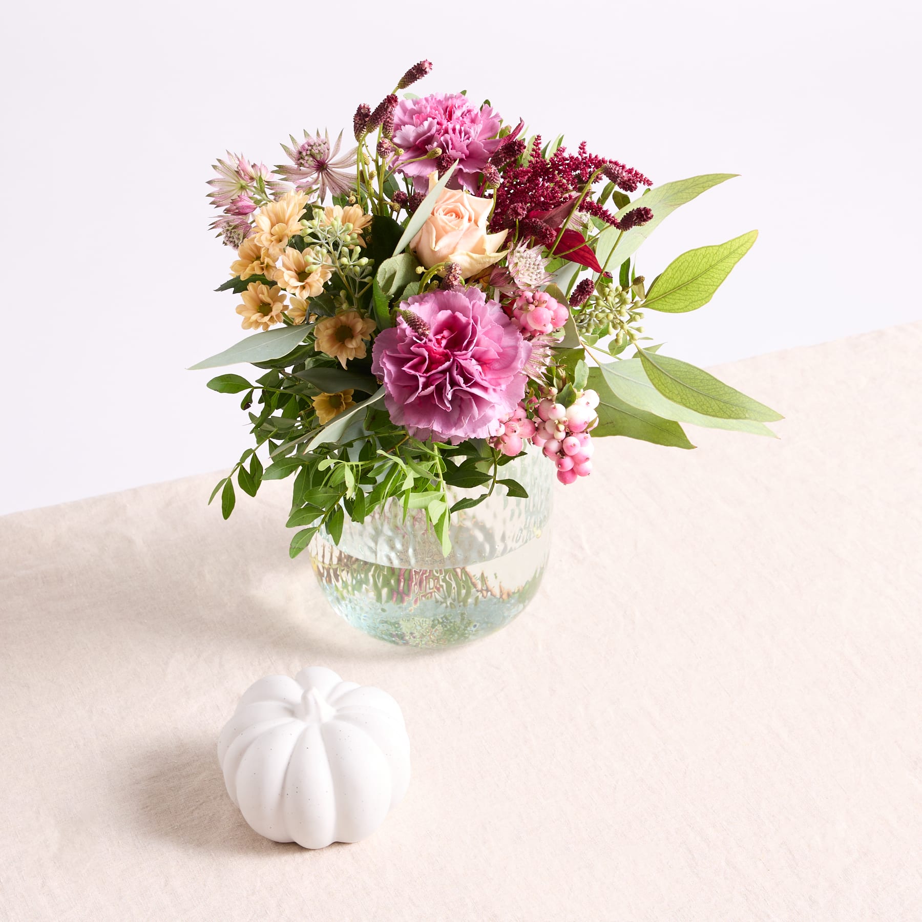 Ein leuchtender Strauß aus rosa Nelken, gelben Gänseblümchen und anderen bunten Blumen in einer Glasvase steht auf einer hellen Oberfläche, begleitet von einem kleinen weißen Kürbis.