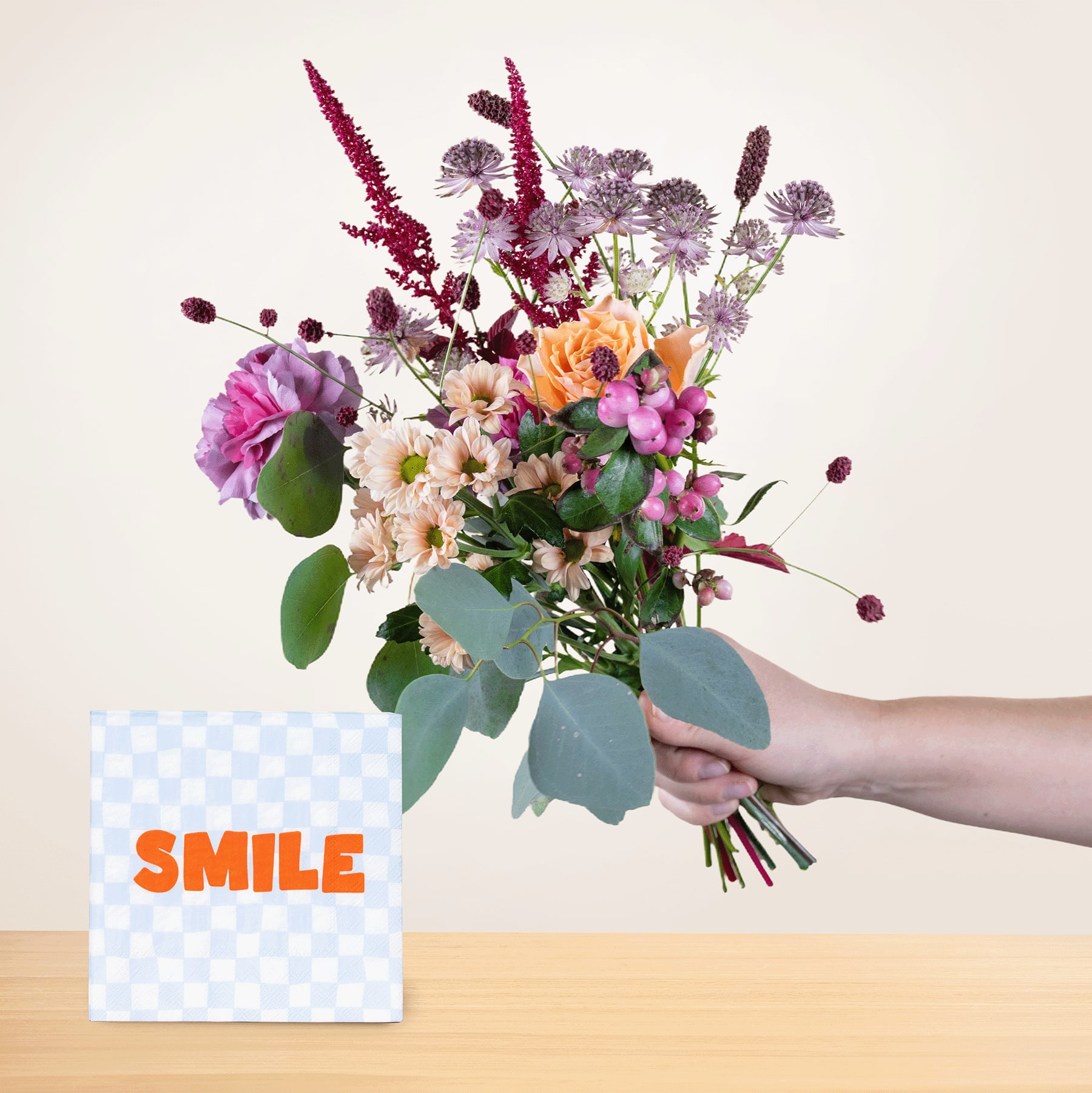 Eine Person hält vor einem schlichten Hintergrund einen leuchtenden Strauß bunter Blumen, darunter Rosen, Gänseblümchen und andere Blüten, in den Händen. Im Vordergrund ist eine Karte mit dem Wort „SMILE“ platziert.
