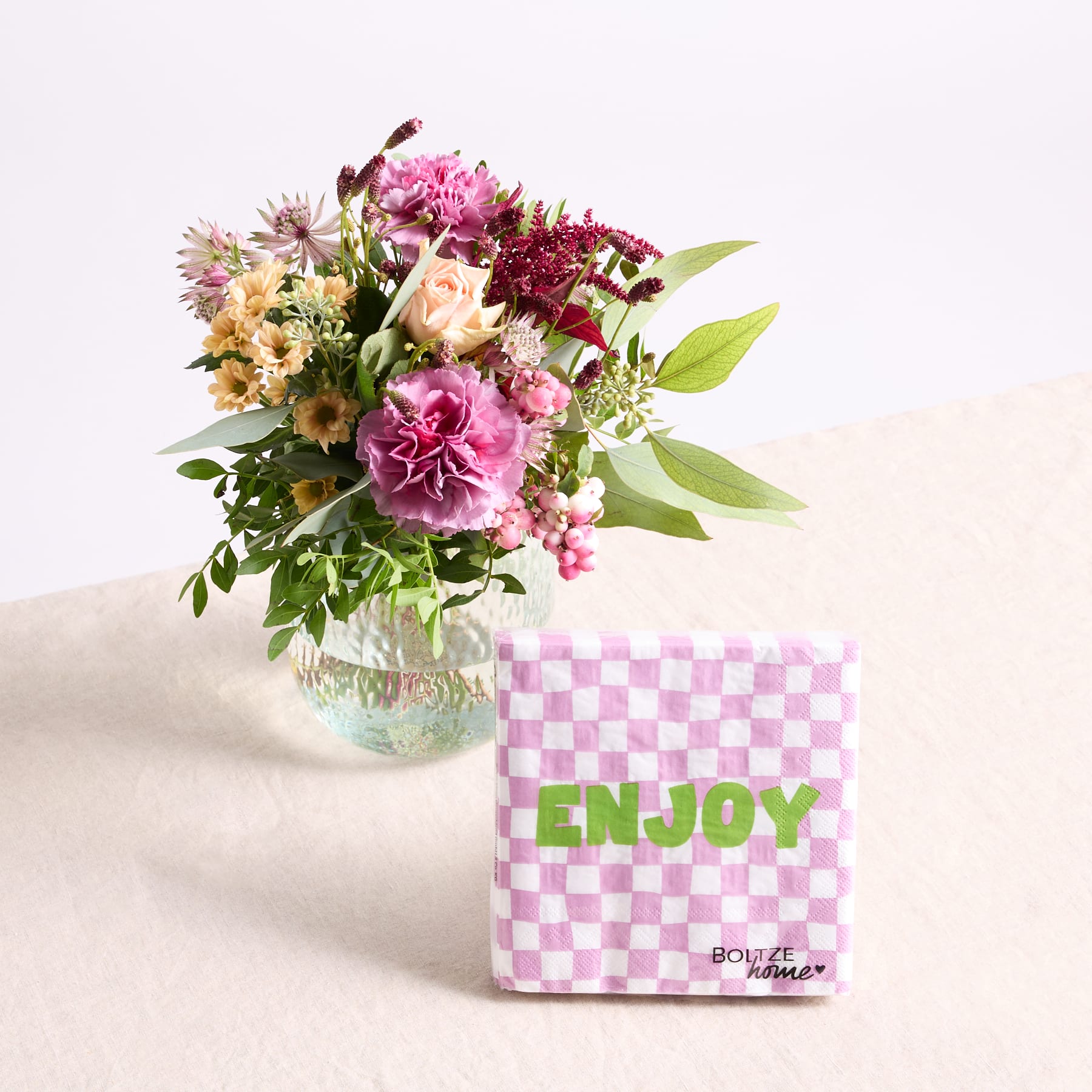 Ein farbenfroher Strauß rosa und weißer Blumen steht in einer Glasvase, begleitet von einer rosa und weiß karierten Schachtel mit dem Aufdruck „ENJOY“, alles vor einem schlichten weißen Hintergrund.