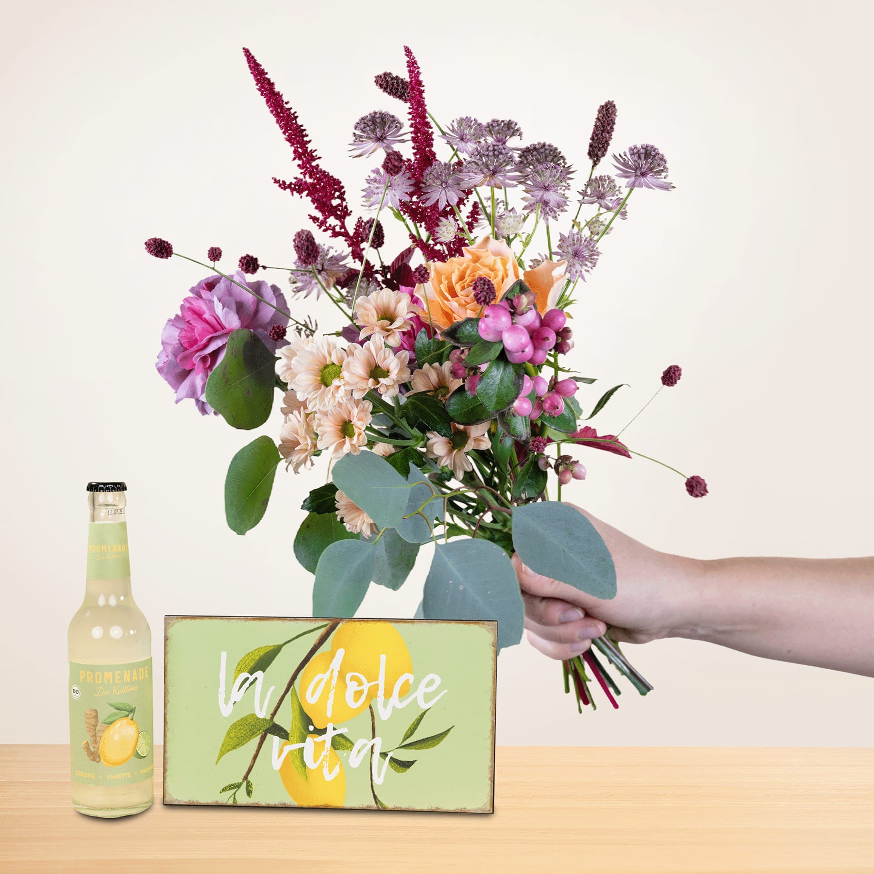 Eine Person hält einen leuchtenden Strauß bunter Blumen, darunter Rosen, Gänseblümchen und andere Blüten, neben einer grünen Schachtel mit der Aufschrift „Dolce Vita“ und einer Flasche eines scheinbar zitronenhaltigen Getränks in den Händen.