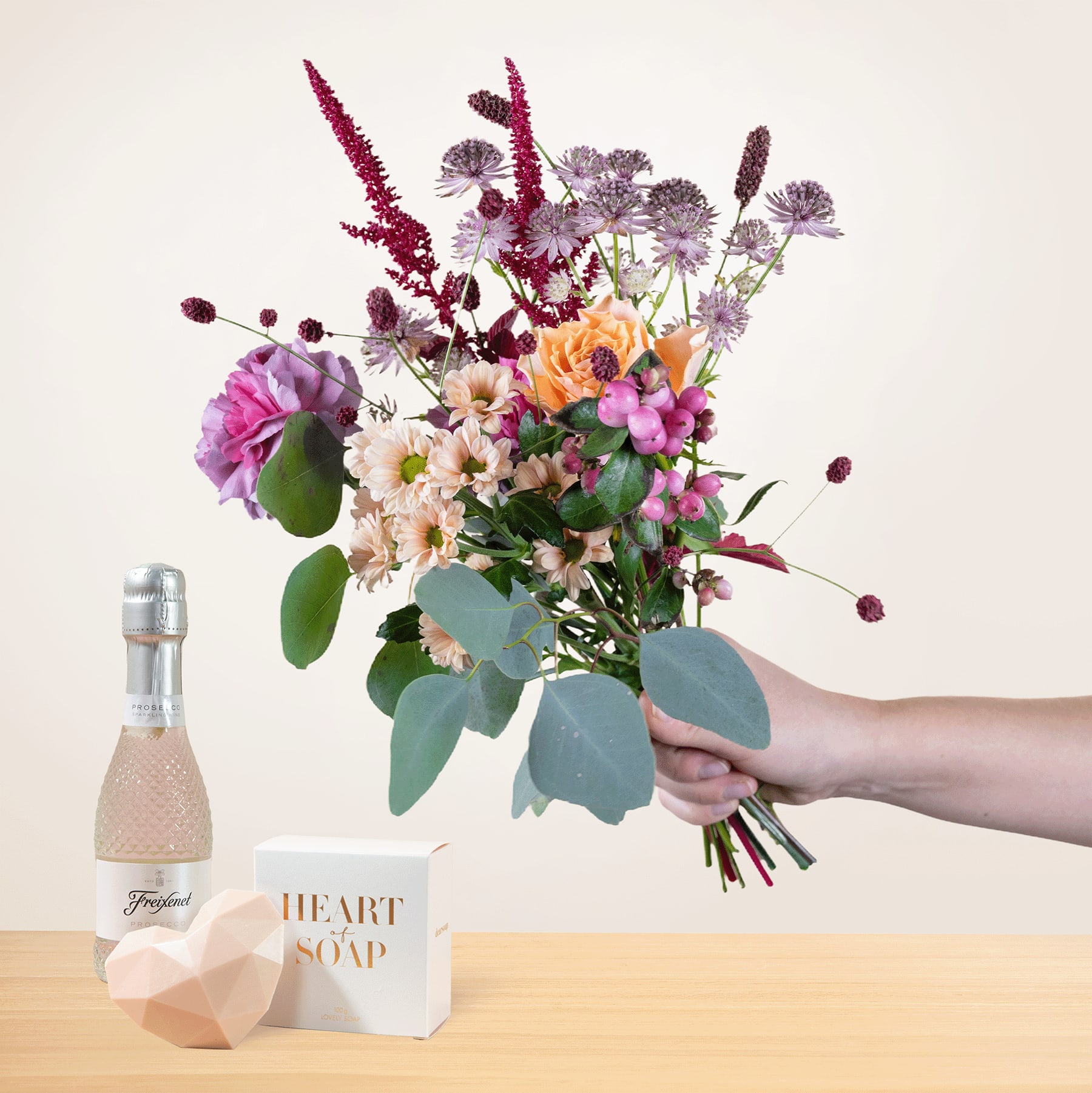 Eine Person hält einen leuchtenden Strauß bunter Blumen, darunter Rosen, Gänseblümchen und andere Blüten, in den Händen, begleitet von einer Flasche herzförmiger Seife und einer Glasflasche auf einer Holzoberfläche.