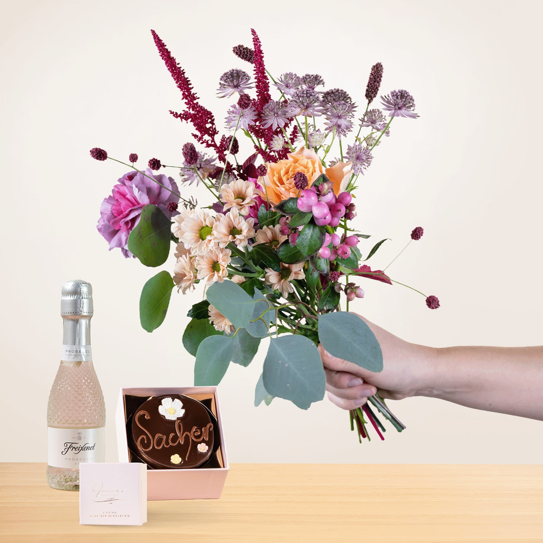 Eine Person hält einen farbenfrohen Strauß bunter Blumen, darunter Rosen, Gänseblümchen und andere Blüten, zusammen mit einer Flasche Sekt und einer kleinen Geschenkbox auf einer Holzoberfläche in den Händen.
