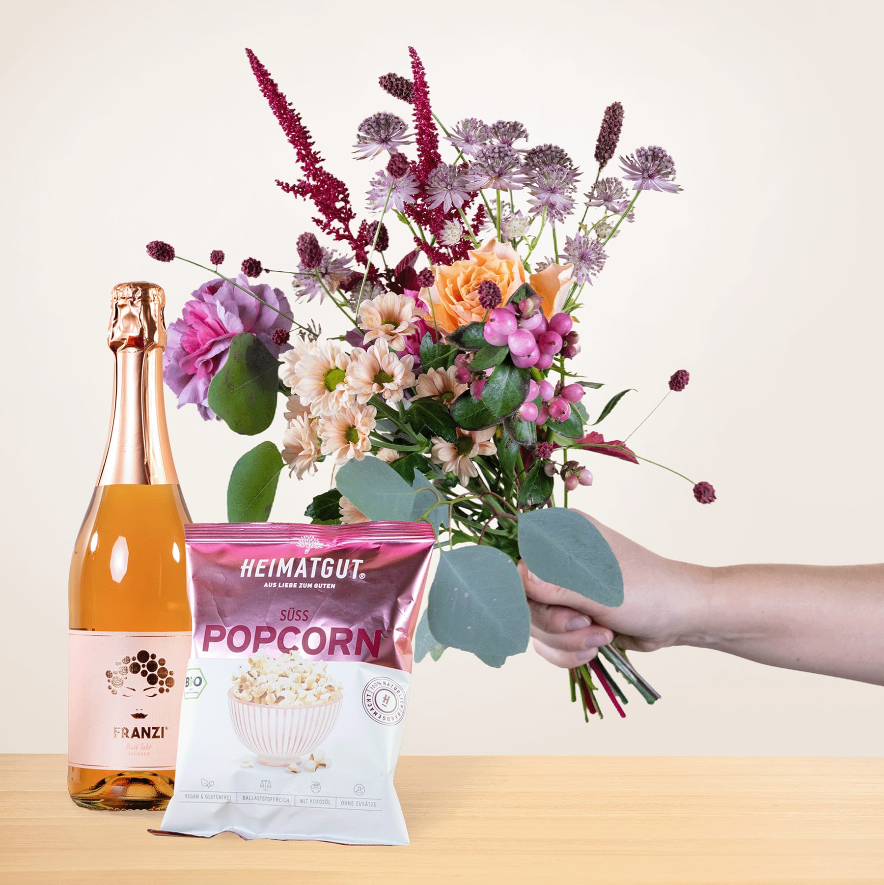 In einer Hand hält man einen leuchtenden Strauß bunter Blumen, darunter Rosa-, Lila- und Orangetöne, zusammen mit einer Flasche Sekt und einer Tüte Popcorn, alles vor einem schlichten Hintergrund.
