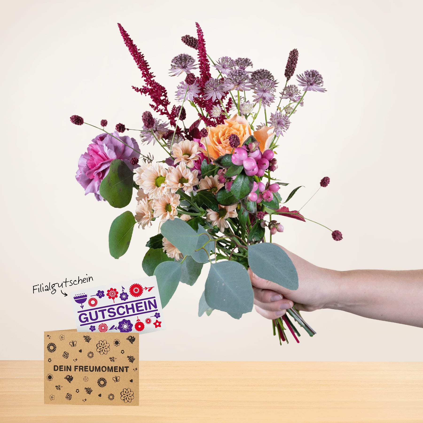 Eine Person hält einen farbenfrohen Strauß bunter Blumen, darunter Rosen, Gänseblümchen und andere Blüten, in der Hand, im Hintergrund ist eine Grußkarte zu sehen.
