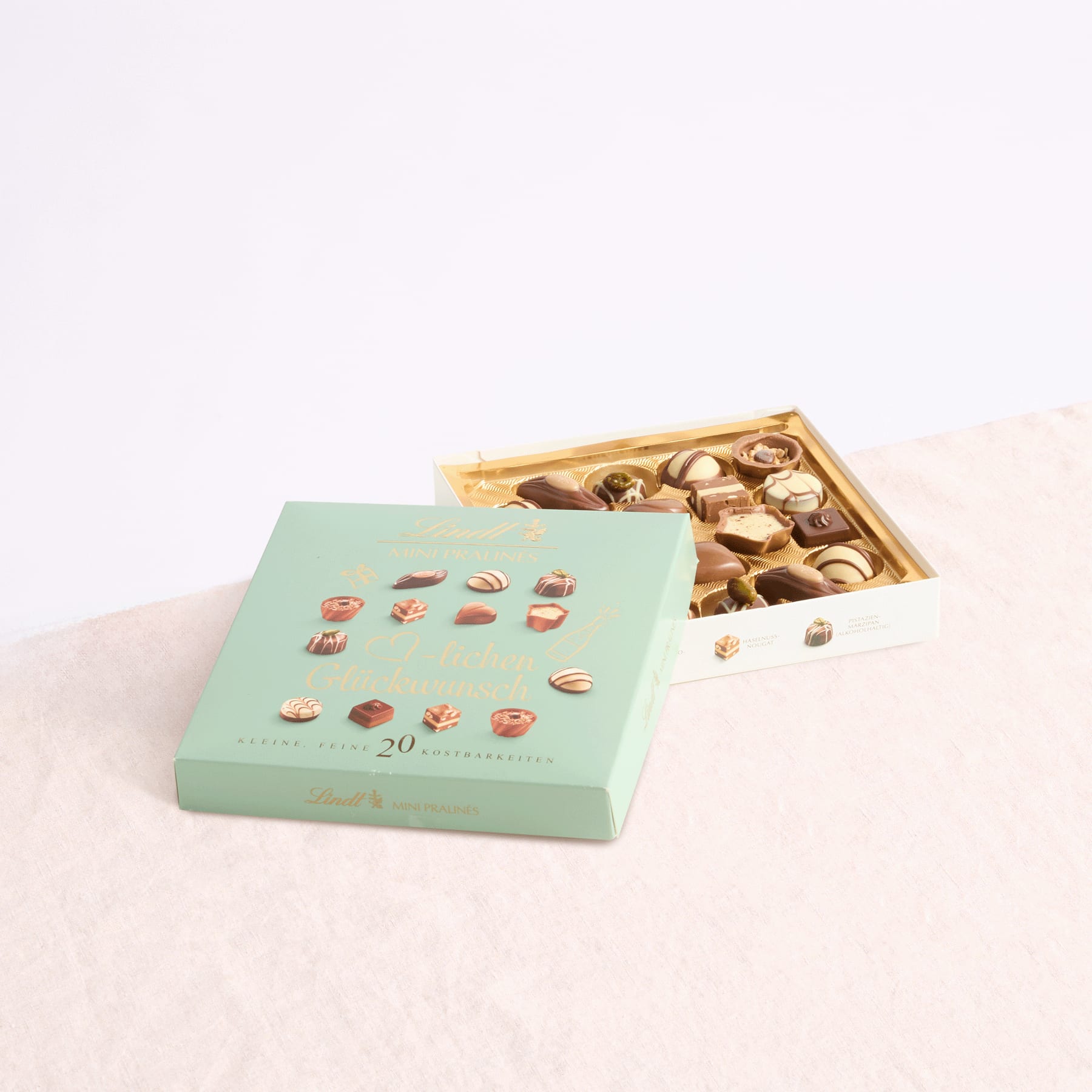 Eine grüne Schachtel mit verschiedenen Pralinen steht neben einer offenen, goldfarbenen Schachtel mit weiteren Pralinen auf einer hellen Oberfläche.