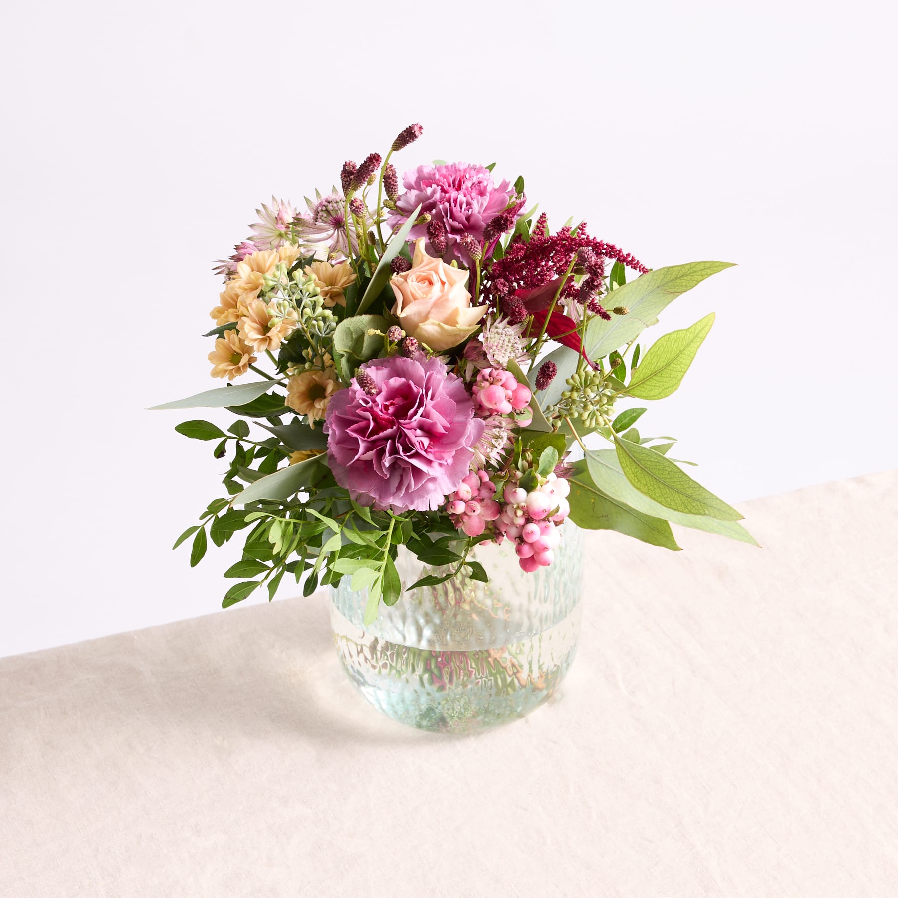 Ein leuchtender Strauß aus rosa Pfingstrosen, gelben Narzissen und anderen bunten Blumen in einer Glasvase steht auf einer hellen Fläche vor einem schlichten Hintergrund.