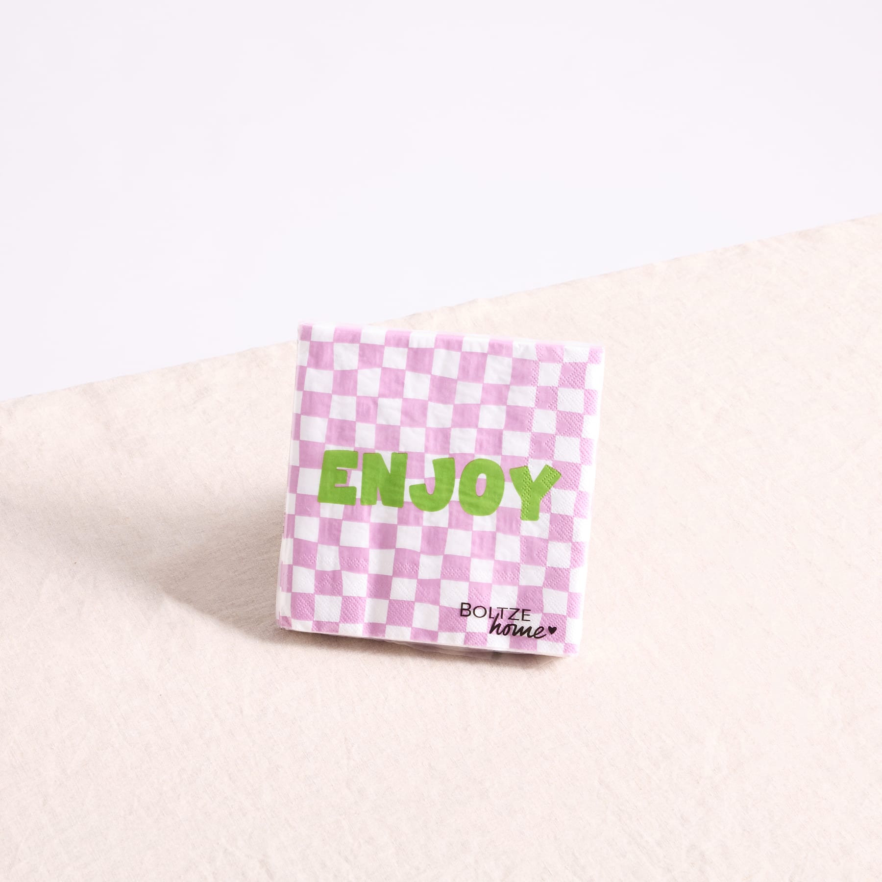 Eine rosa-weiß karierte Papierserviette mit dem in grüner Schrift aufgedruckten Wort „ENJOY“ liegt auf einer hellen Oberfläche.