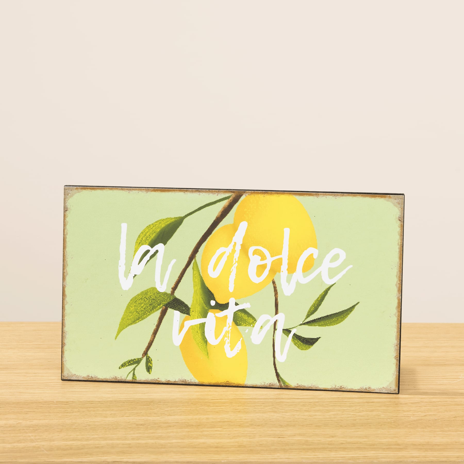 Ein Holzschild mit dem Text „La dolce vita“ auf grün-gelbem Hintergrund, mit Blättern und einer Zitrone.