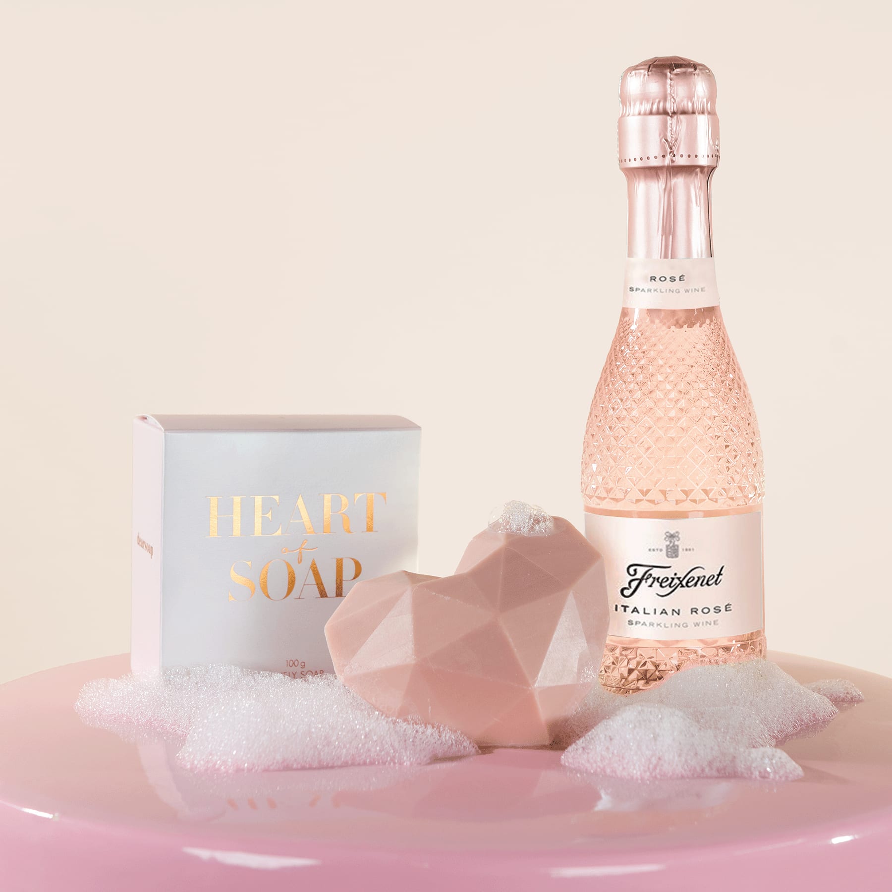 Eine rosa getönte Glasflasche Sekt, eine weiße Schachtel mit der Aufschrift „Luxusseife“ und ein rosafarbener, geometrisch geformter Gegenstand stehen auf einer weichen, flauschigen rosafarbenen Oberfläche.
