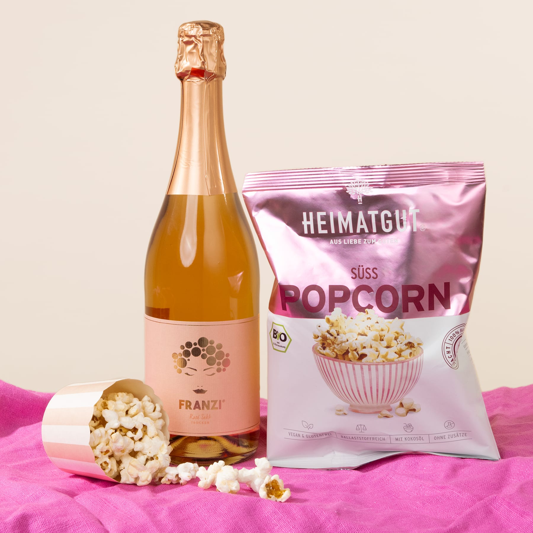Auf einem rosa Stoffhintergrund sind eine Flasche Sekt, eine Tüte süßes Popcorn und verstreute Popcornkörner arrangiert.