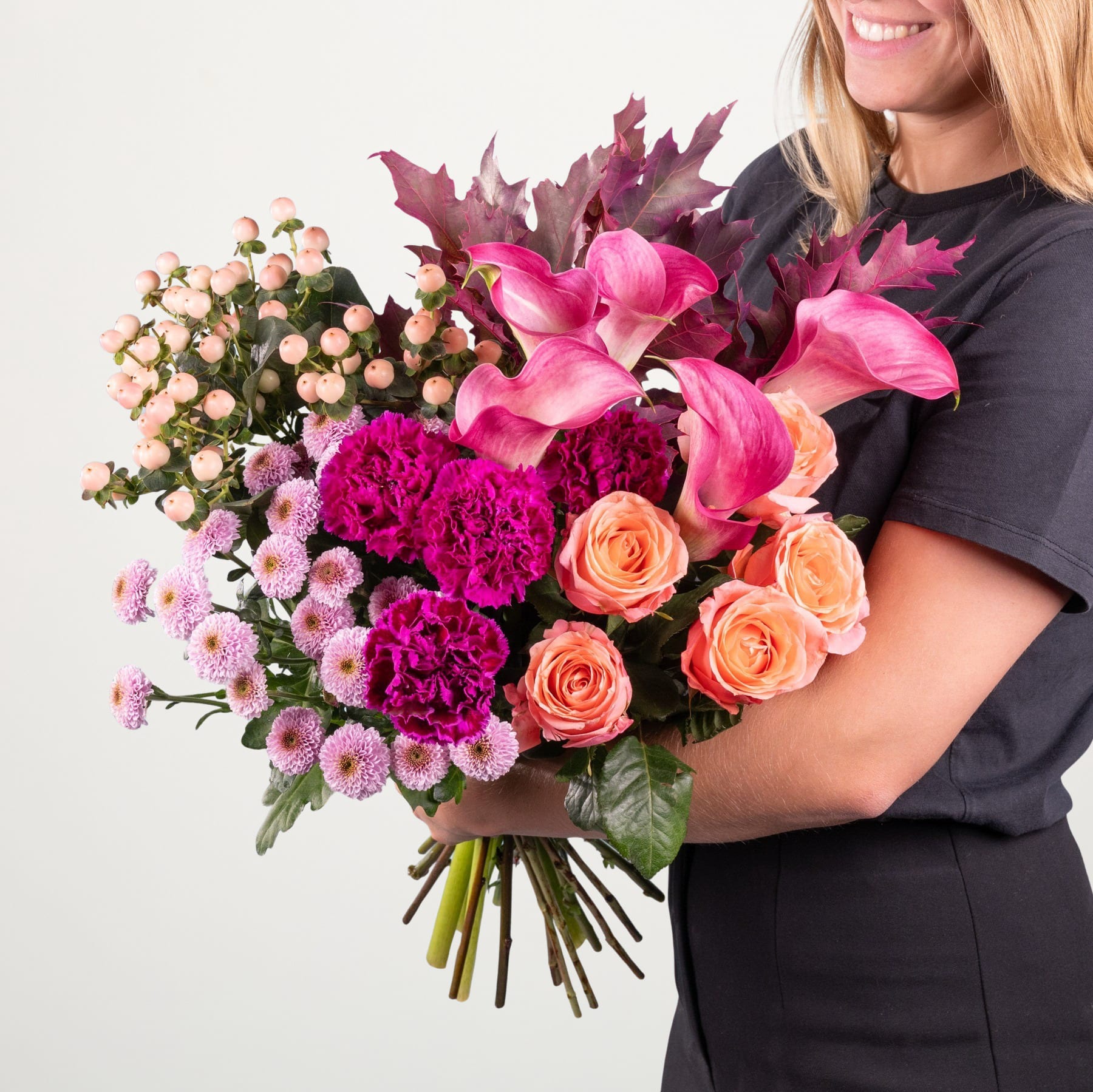 Ein leuchtender Strauß bunter Blumen, darunter rosa Rosen, Nelken und Chrysanthemen, wird von einer Person gehalten, die ein schwarzes Hemd vor einem schlichten weißen Hintergrund trägt.