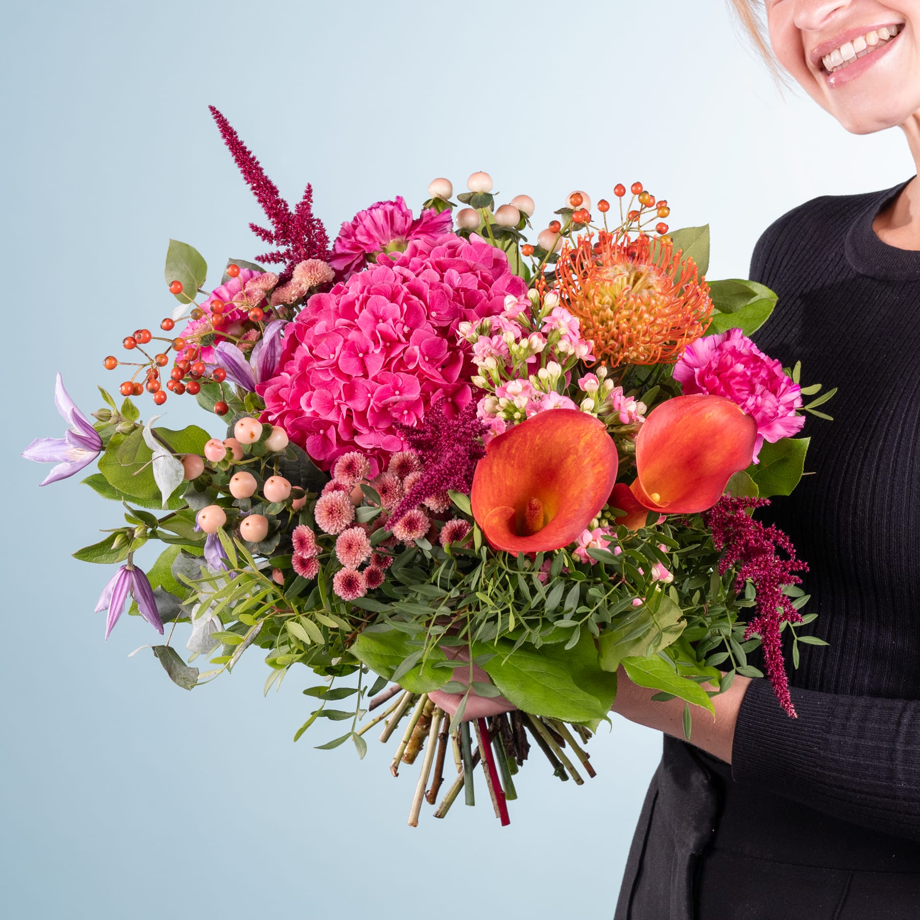 Ein leuchtender Strauß bunter Blumen, darunter rosa Hortensien, orangefarbene Proteen und verschiedene andere Blüten, gehalten von einer Person in einem schwarzen Outfit vor einem schlichten Hintergrund.