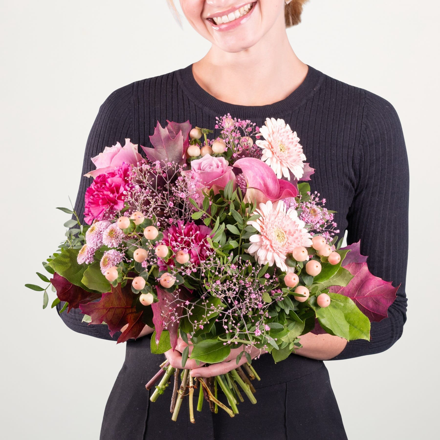 Eine lächelnde Person in einem schwarzen Pullover hält einen farbenfrohen Strauß rosa, weißer und violetter Blumen vor einem schlichten Hintergrund.