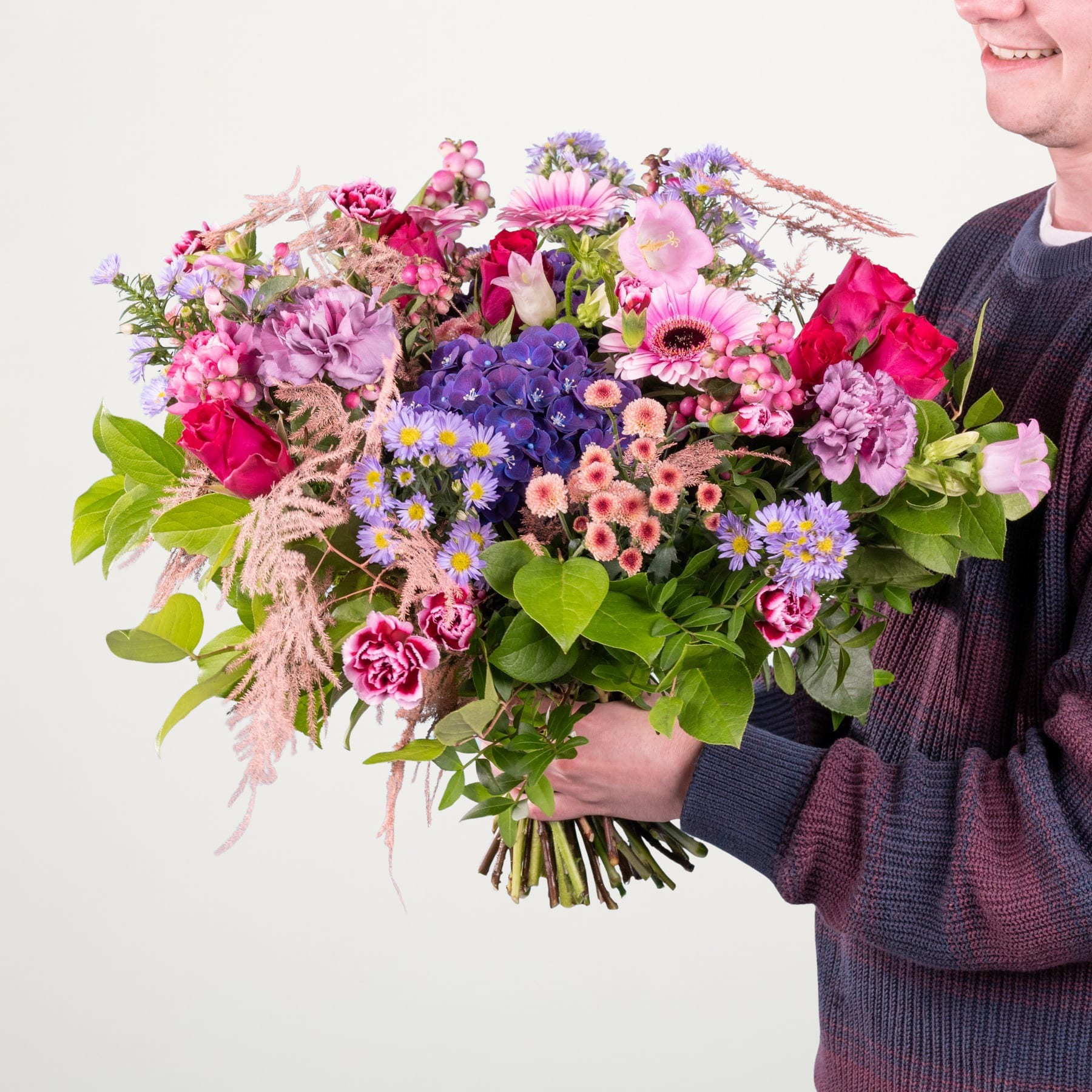 Ein leuchtender Strauß bunter Blumen, darunter rosa Nelken, violette Hortensien und verschiedene andere Blüten, wird von einer Person in einem dunklen Pullover vor einem schlichten weißen Hintergrund gehalten.