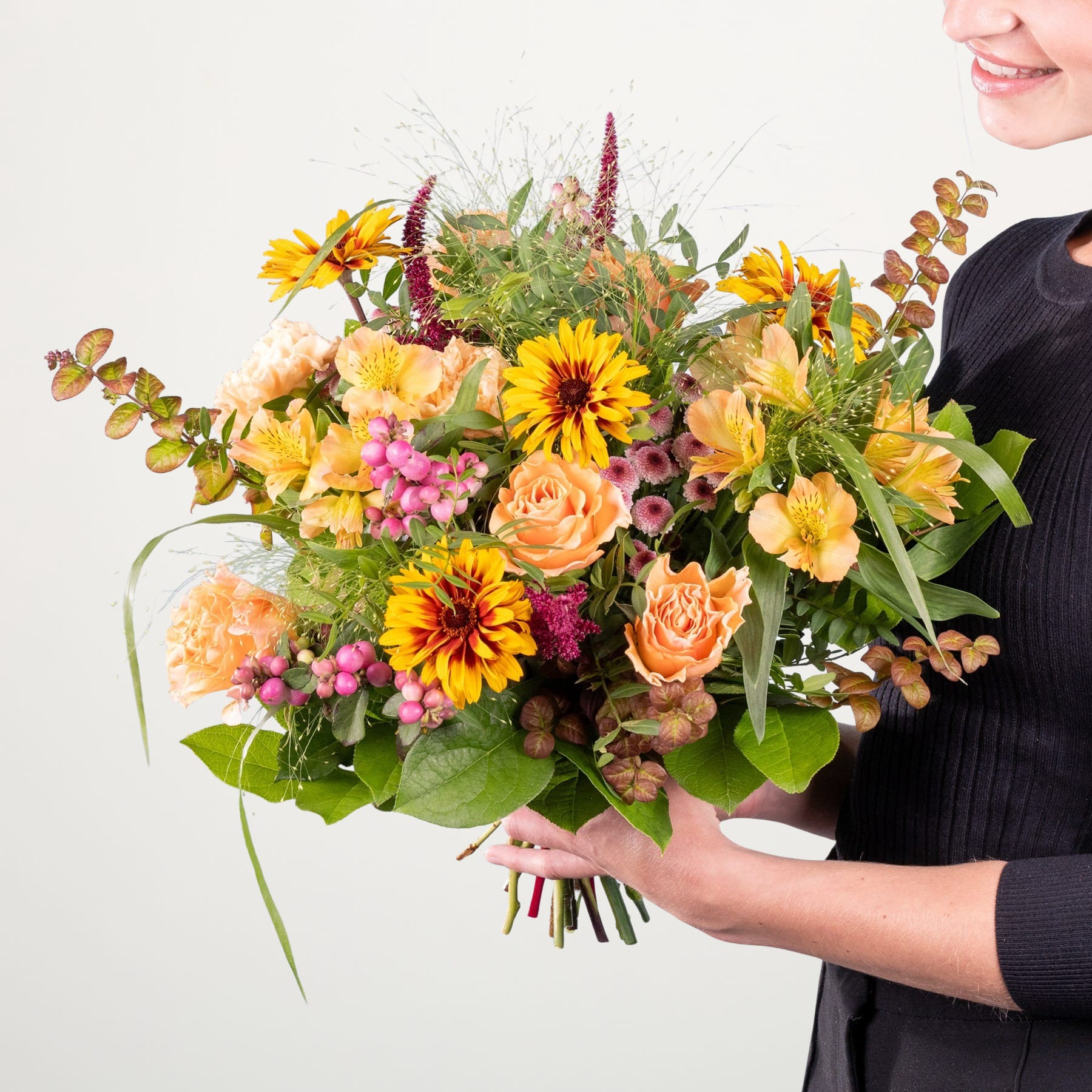 Ein leuchtender Strauß aus gelben Sonnenblumen, orangefarbenen Rosen und verschiedenen anderen bunten Blumen wird von einer Person in einem schwarzen Outfit vor einem schlichten weißen Hintergrund gehalten.