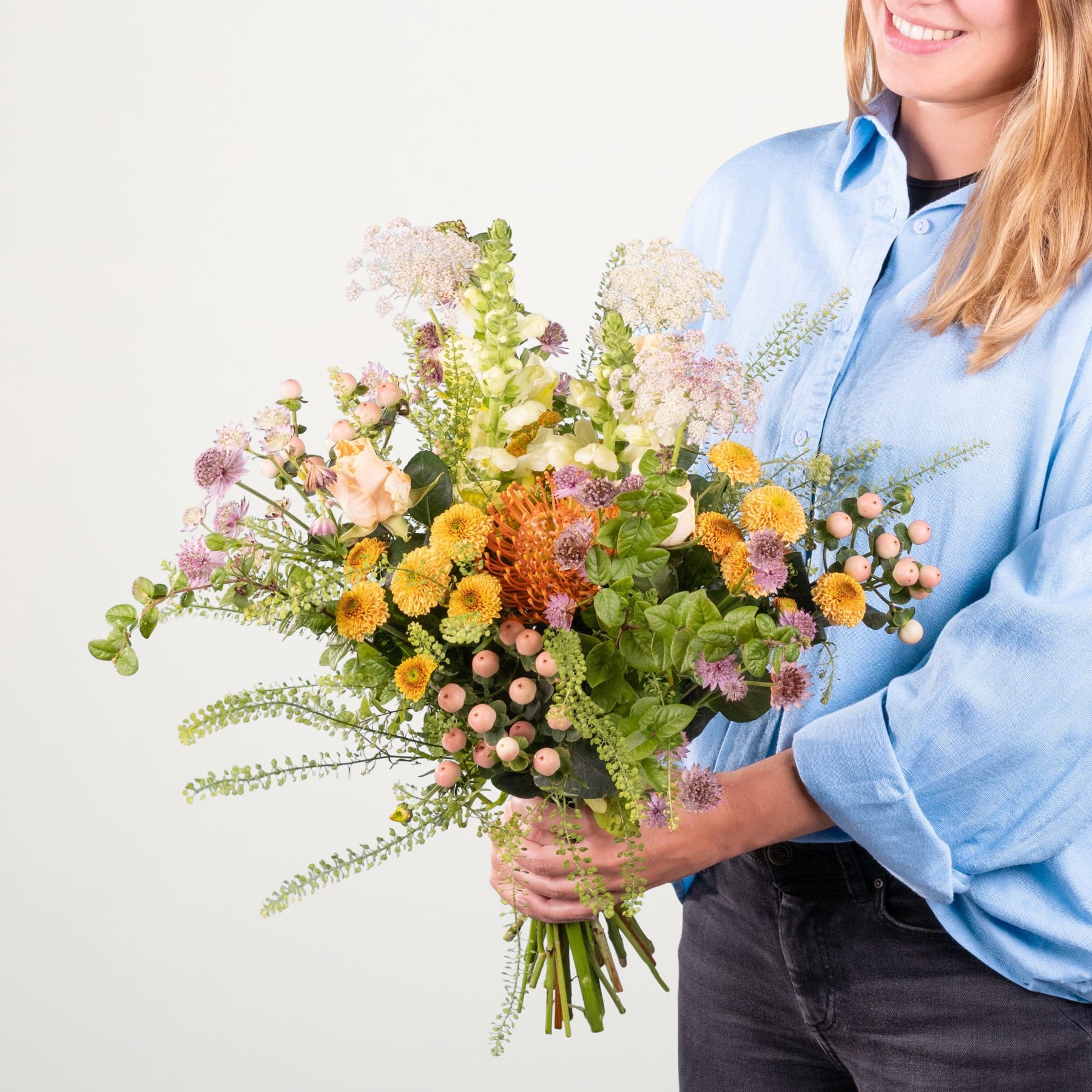 Eine Person in einem blauen Hemd hält vor einem schlichten weißen Hintergrund einen leuchtenden Strauß bunter Blumen, darunter gelbe, orange und rosa Blüten.