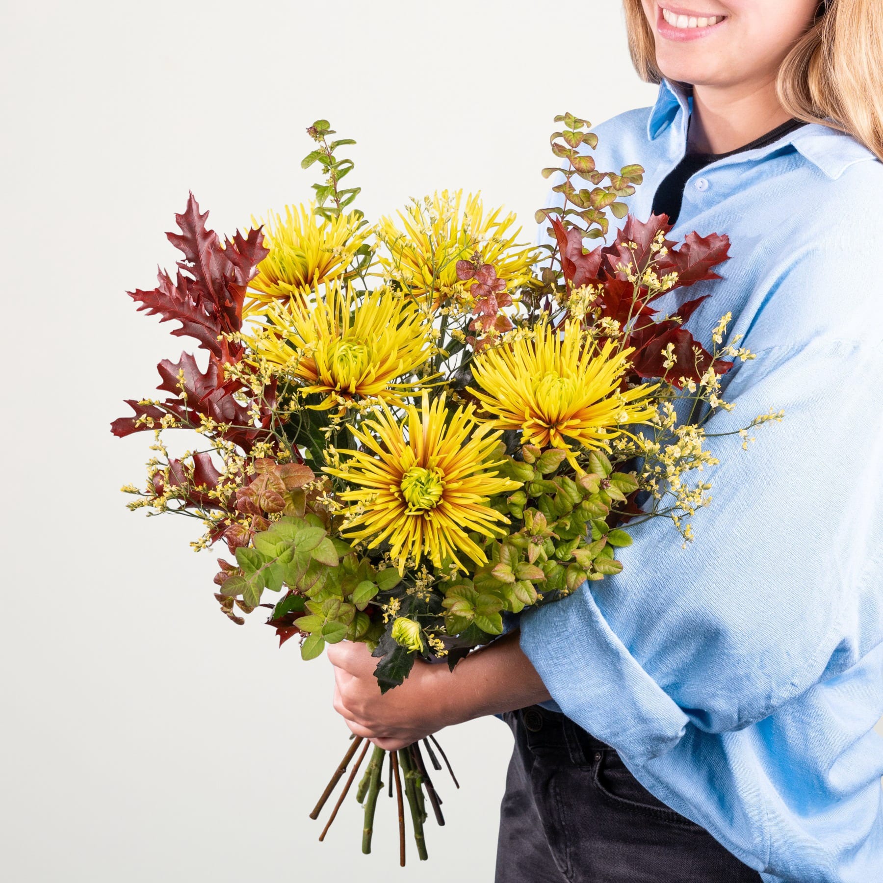 Ein Strauß leuchtend gelber Chrysanthemen und roter Herbstblätter wird von einer lächelnden Frau in einem blauen Hemd vor einem schlichten weißen Hintergrund gehalten.
