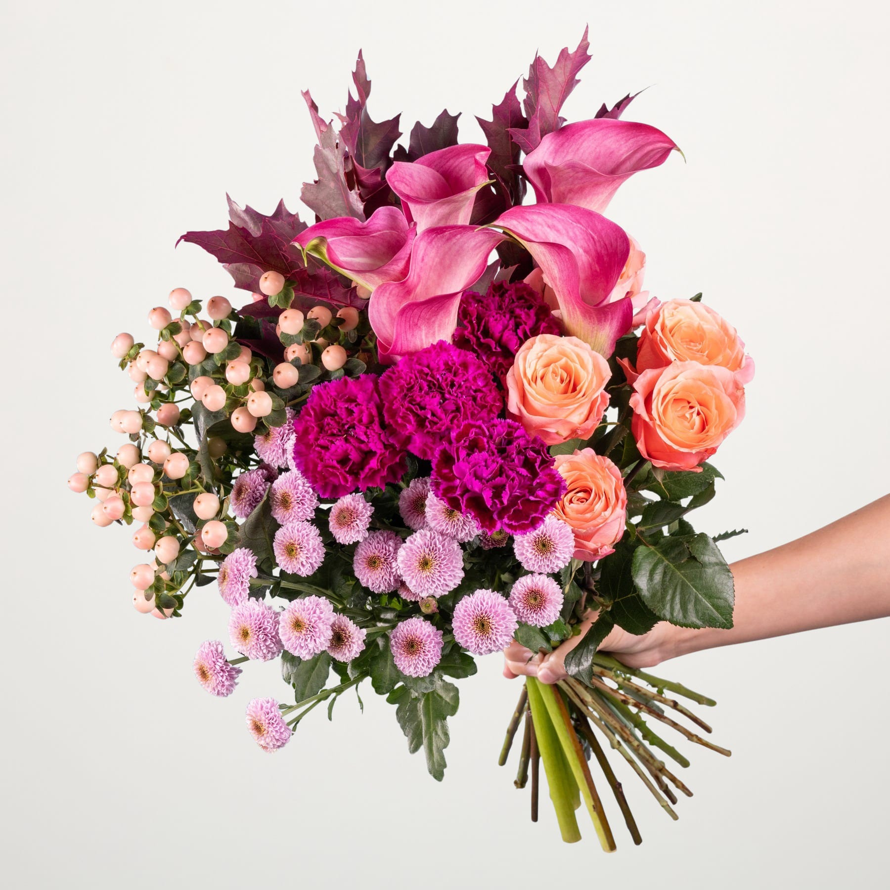 Ein farbenfroher Strauß verschiedener rosa und orangefarbener Blumen, darunter Rosen, Nelken und Chrysanthemen, wird vor einem schlichten weißen Hintergrund in den Händen einer Person gehalten.